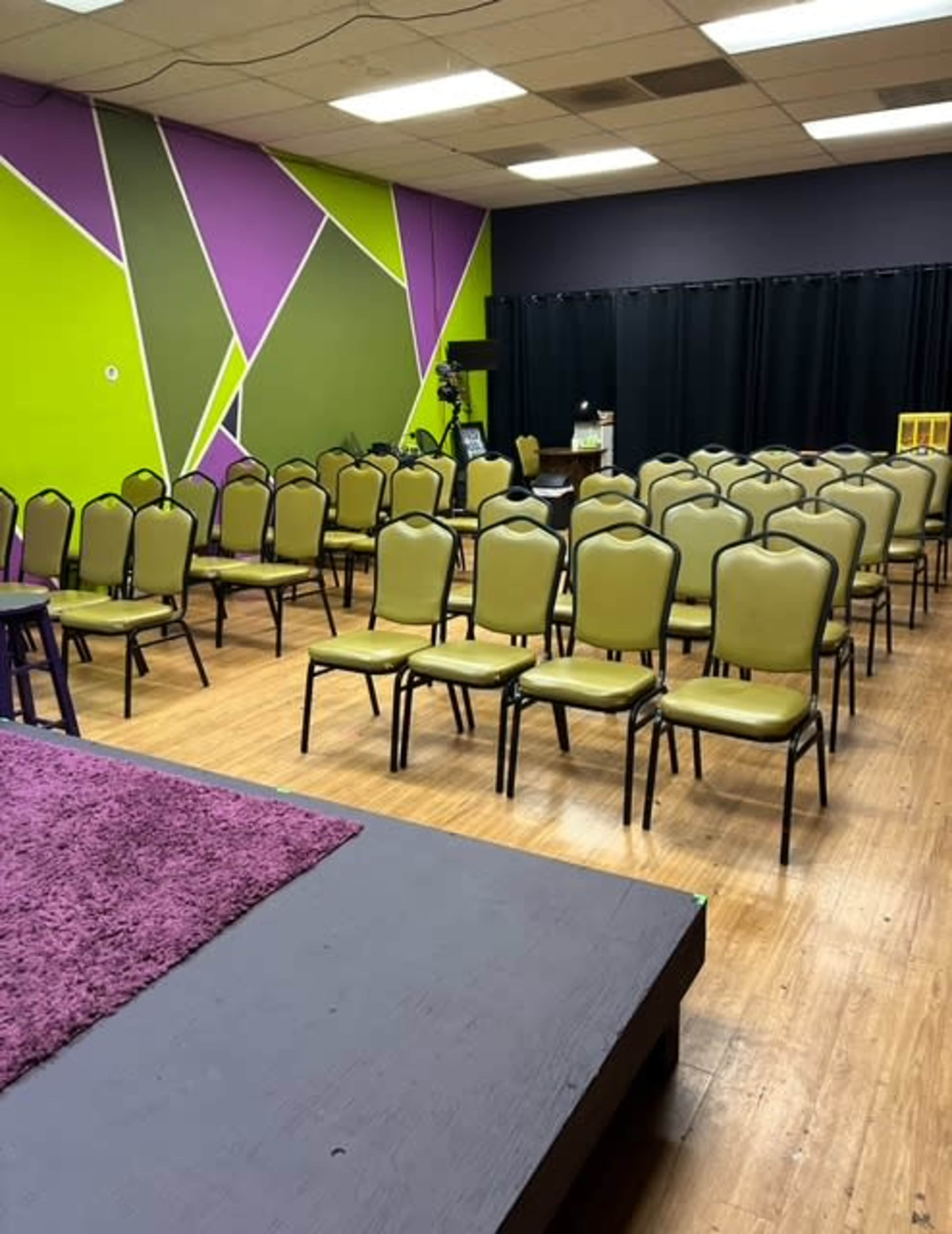 The image shows a room set up with numerous green chairs arranged in rows facing a stage area, with colorful geometric patterns on the walls.