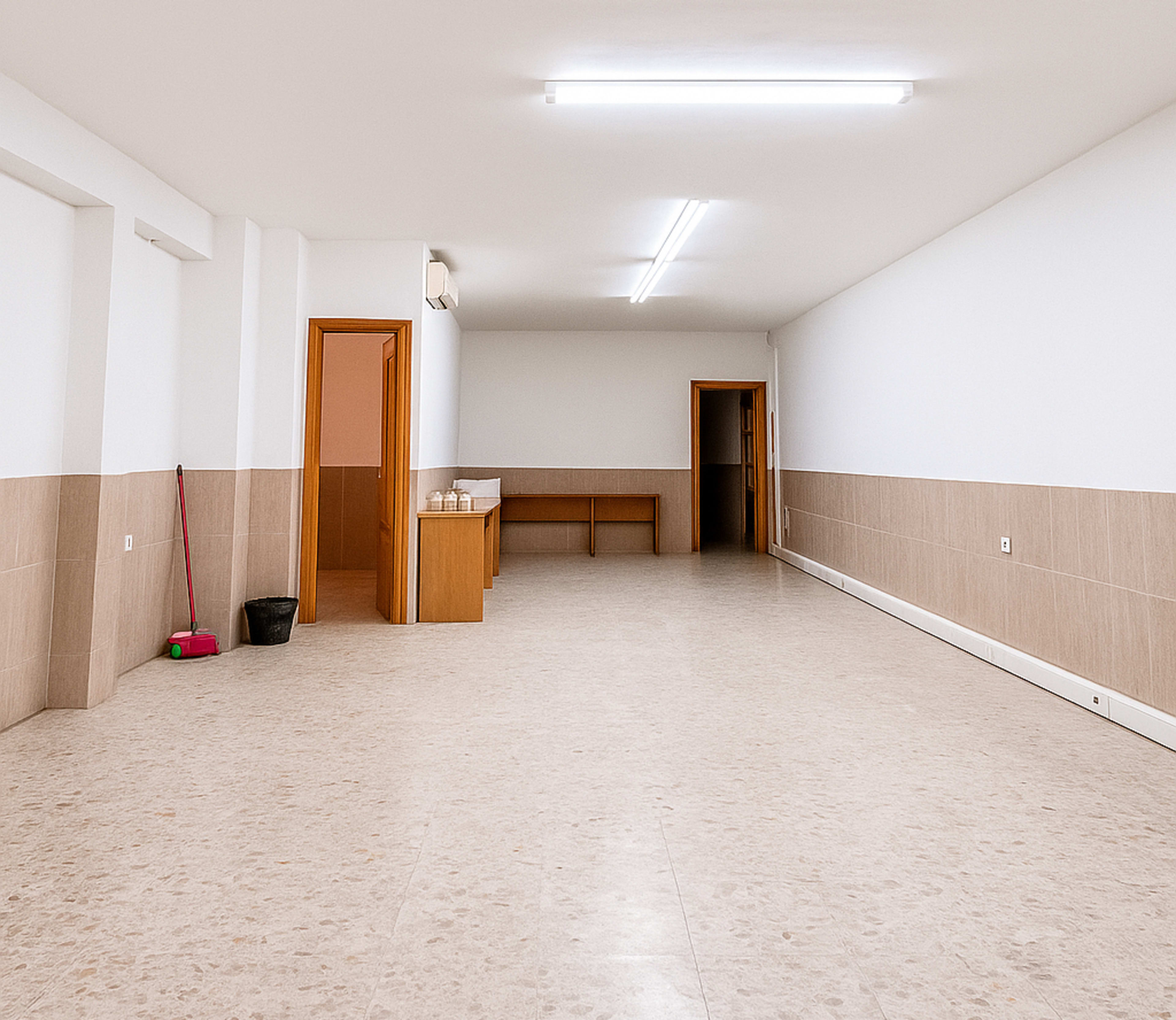 The image shows a spacious, empty room with a light-colored floor, white walls, and a wooden counter along one side.