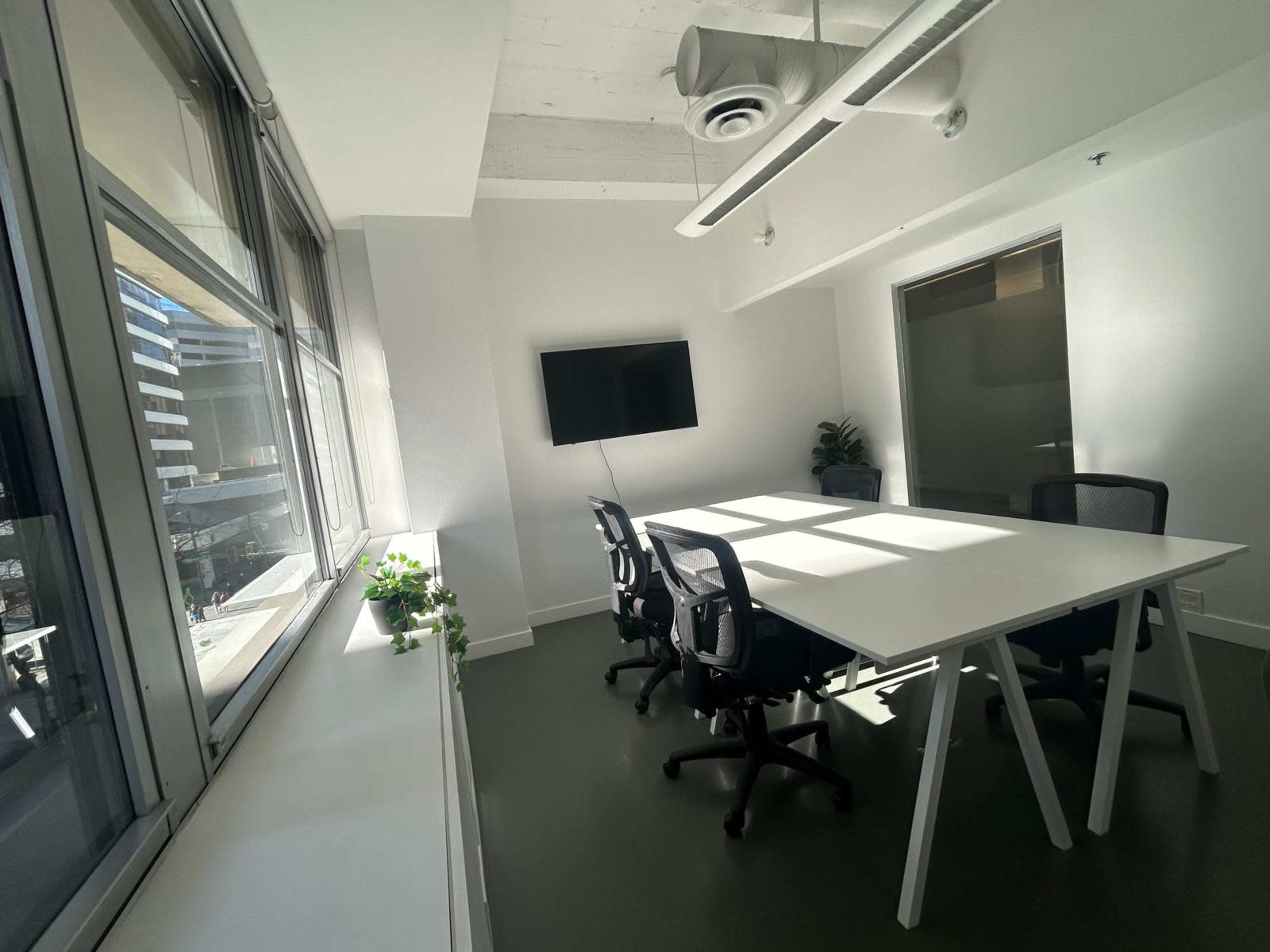 A modern conference room features a sleek white table surrounded by black office chairs, with large windows providing natural light.