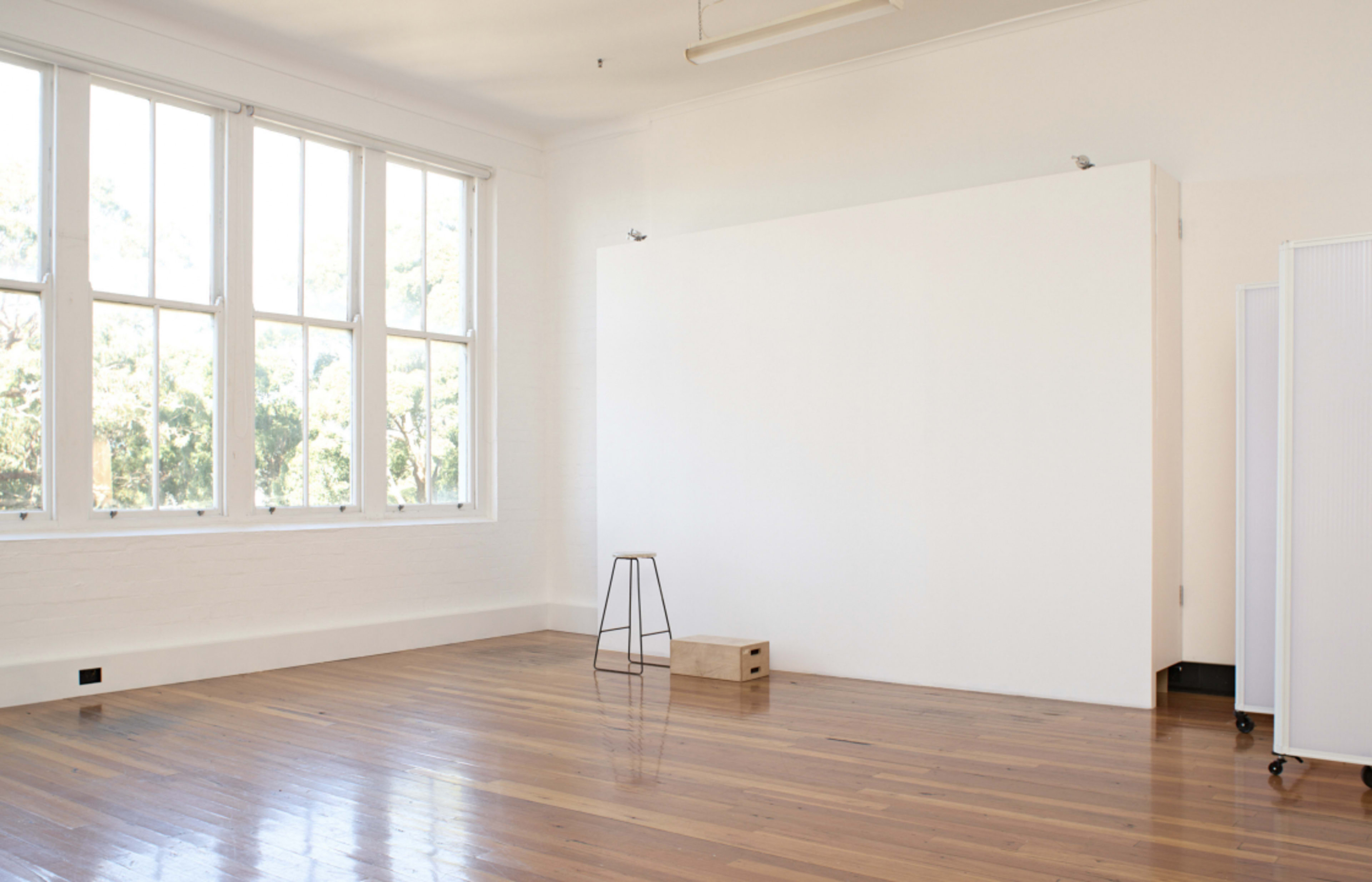 A spacious room with polished wooden floors contains a tall stool and a small wooden box against a plain white wall, illuminated by sunlight through large windows.