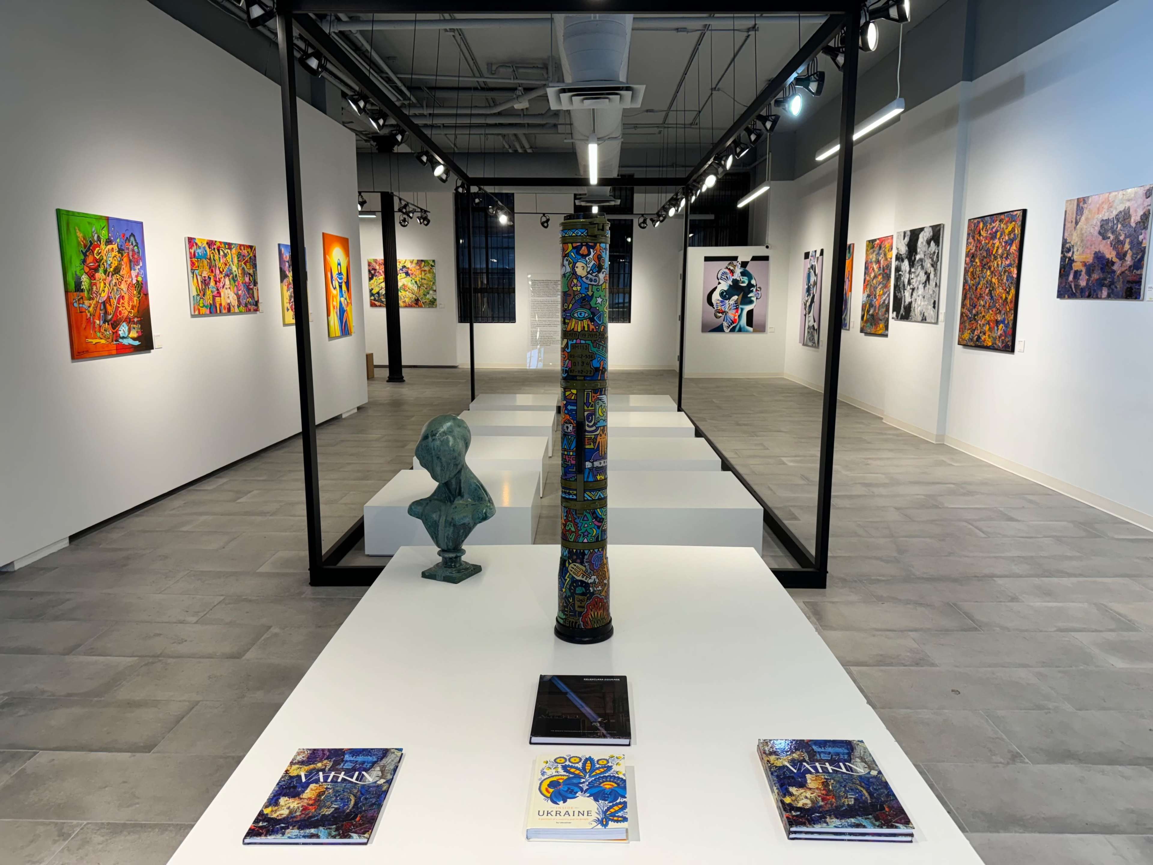 An art gallery featuring colorful paintings on the walls, a tall decorative column in the center, and sculptures displayed on a white platform.