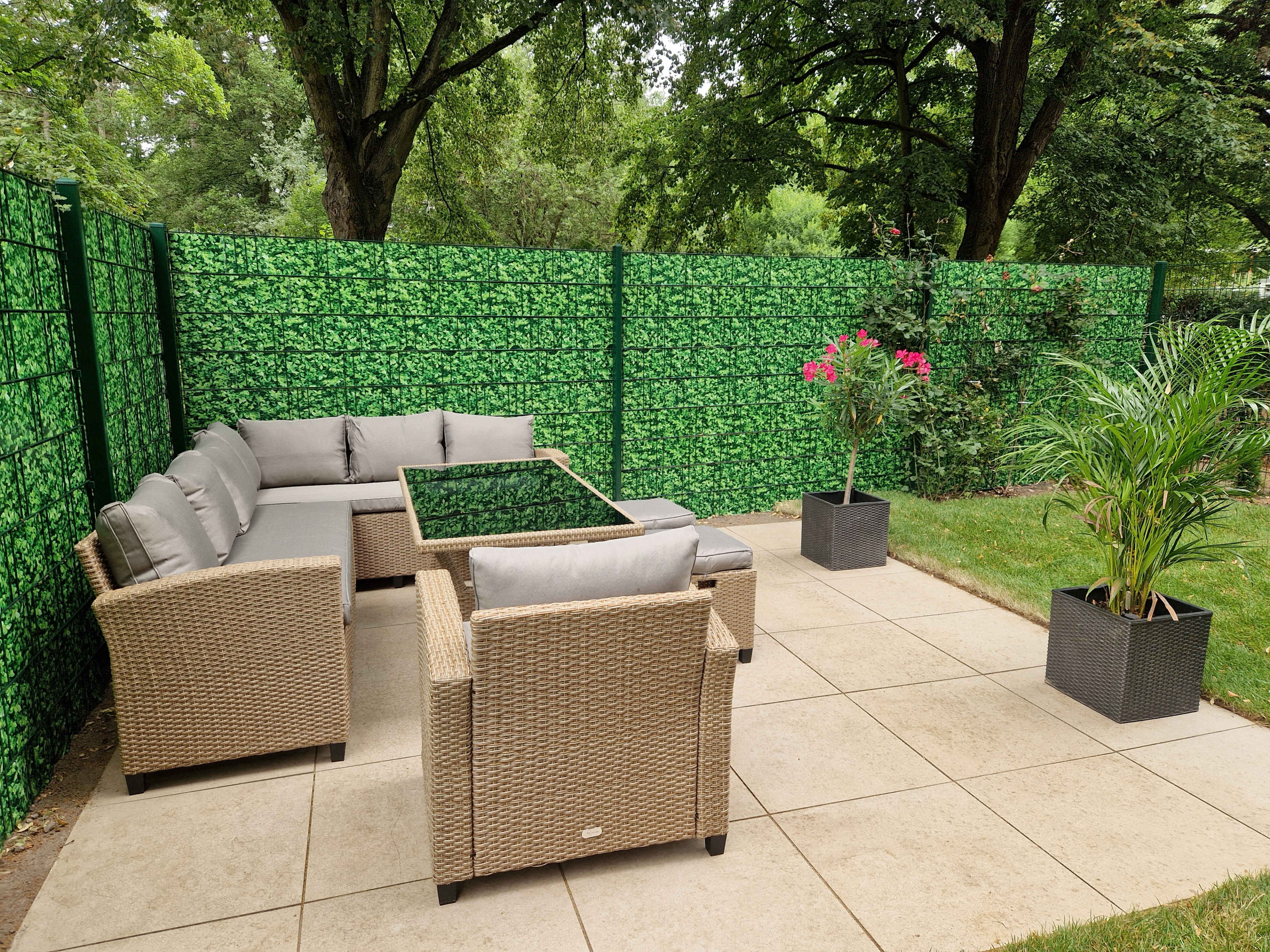 The image shows a landscaped outdoor seating area with a wicker sofa set, glass coffee table, and potted plants surrounded by a green privacy fence.