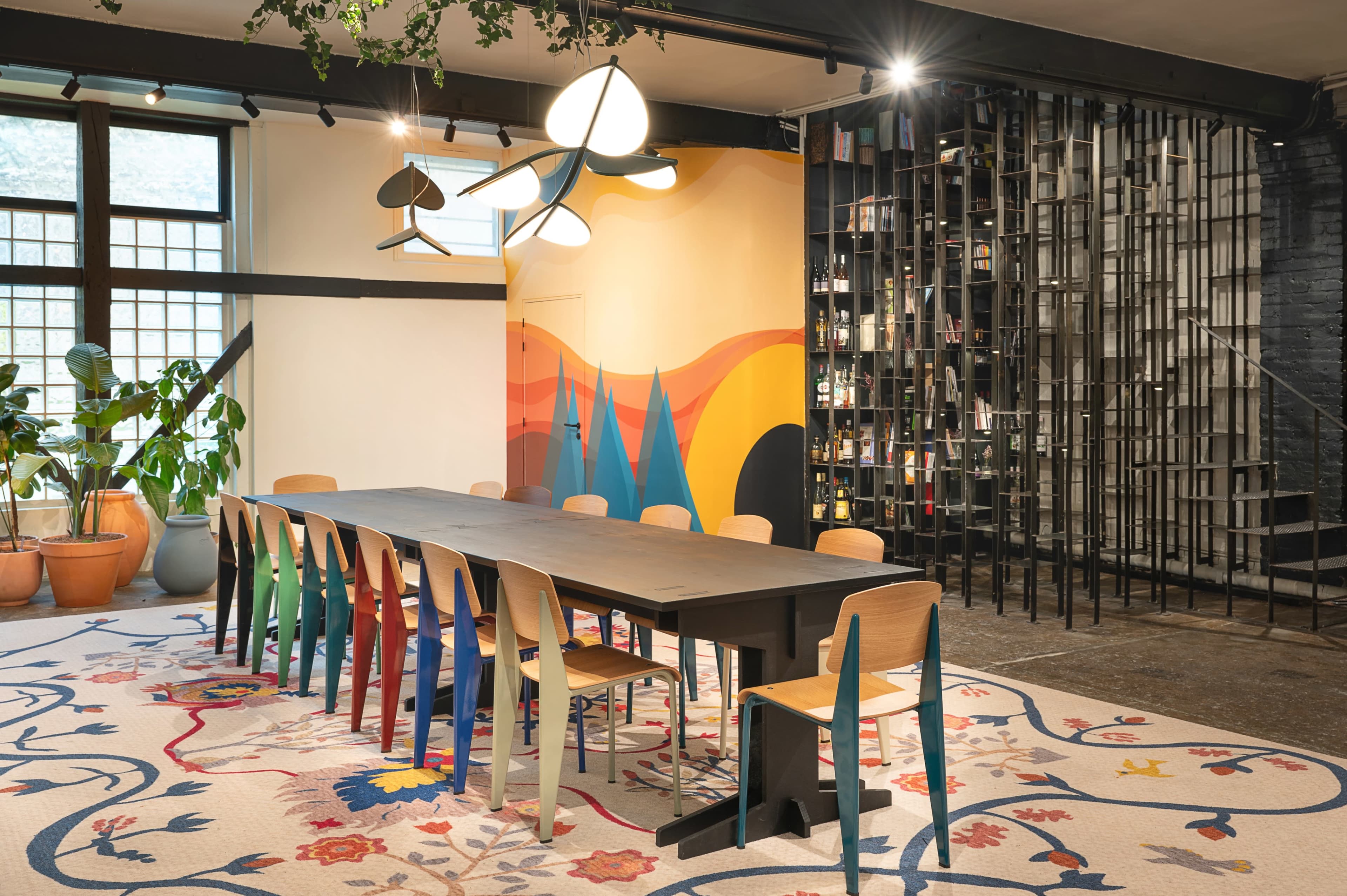 The image depicts a modern dining room featuring a long, dark wooden table surrounded by colorful chairs, a vibrant mural on the wall, and a unique wine storage area.