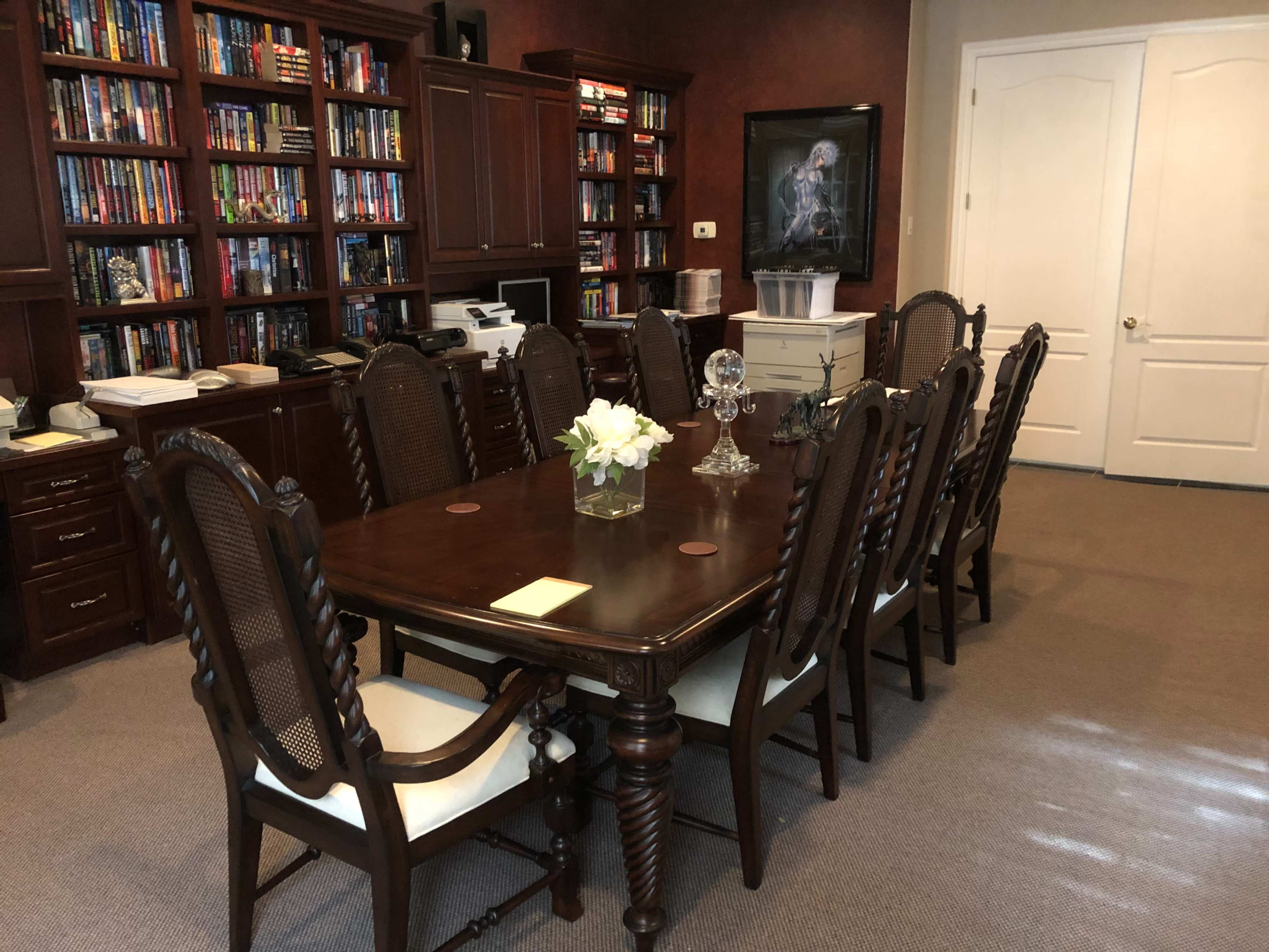 The image shows a well-furnished conference room with a large wooden table surrounded by chairs, set against a backdrop of bookshelves filled with various books.