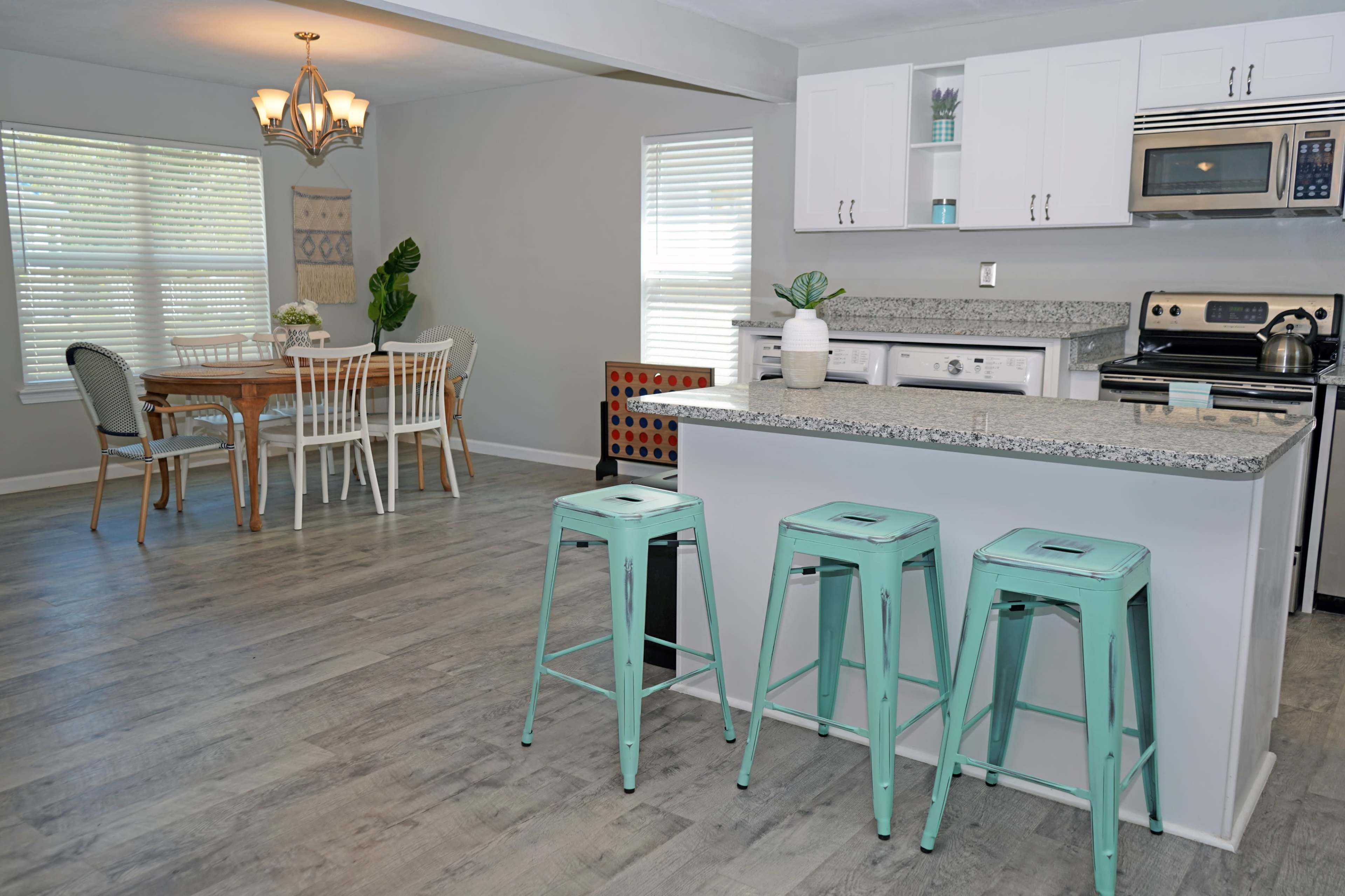 A modern kitchen and dining area with a granite countertop island, three teal stools, a wooden dining table, and various light-colored furnishings.