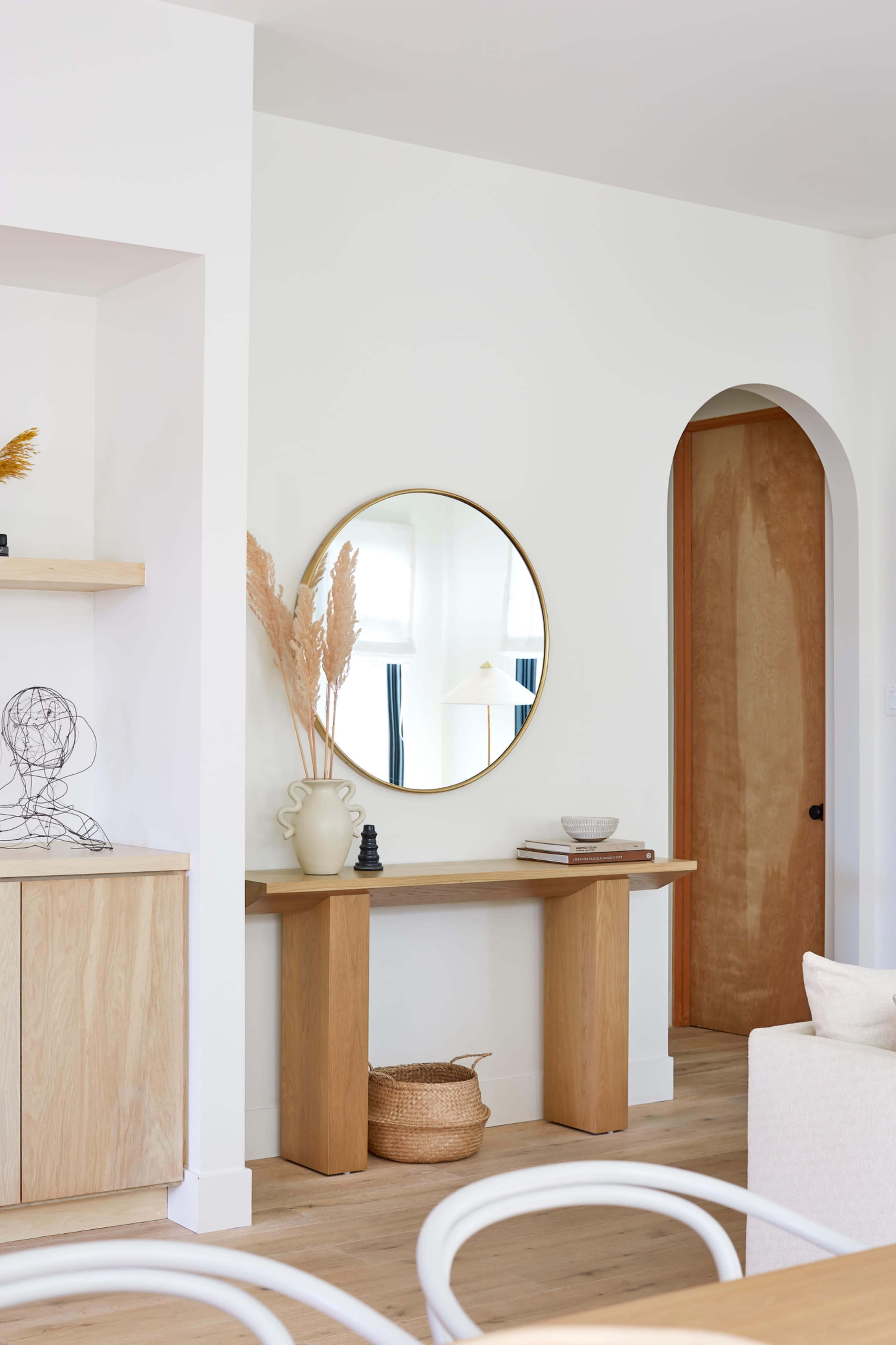 A wooden console table with a circular mirror above it is positioned against a white wall, with a doorway leading to another room visible in the background.
