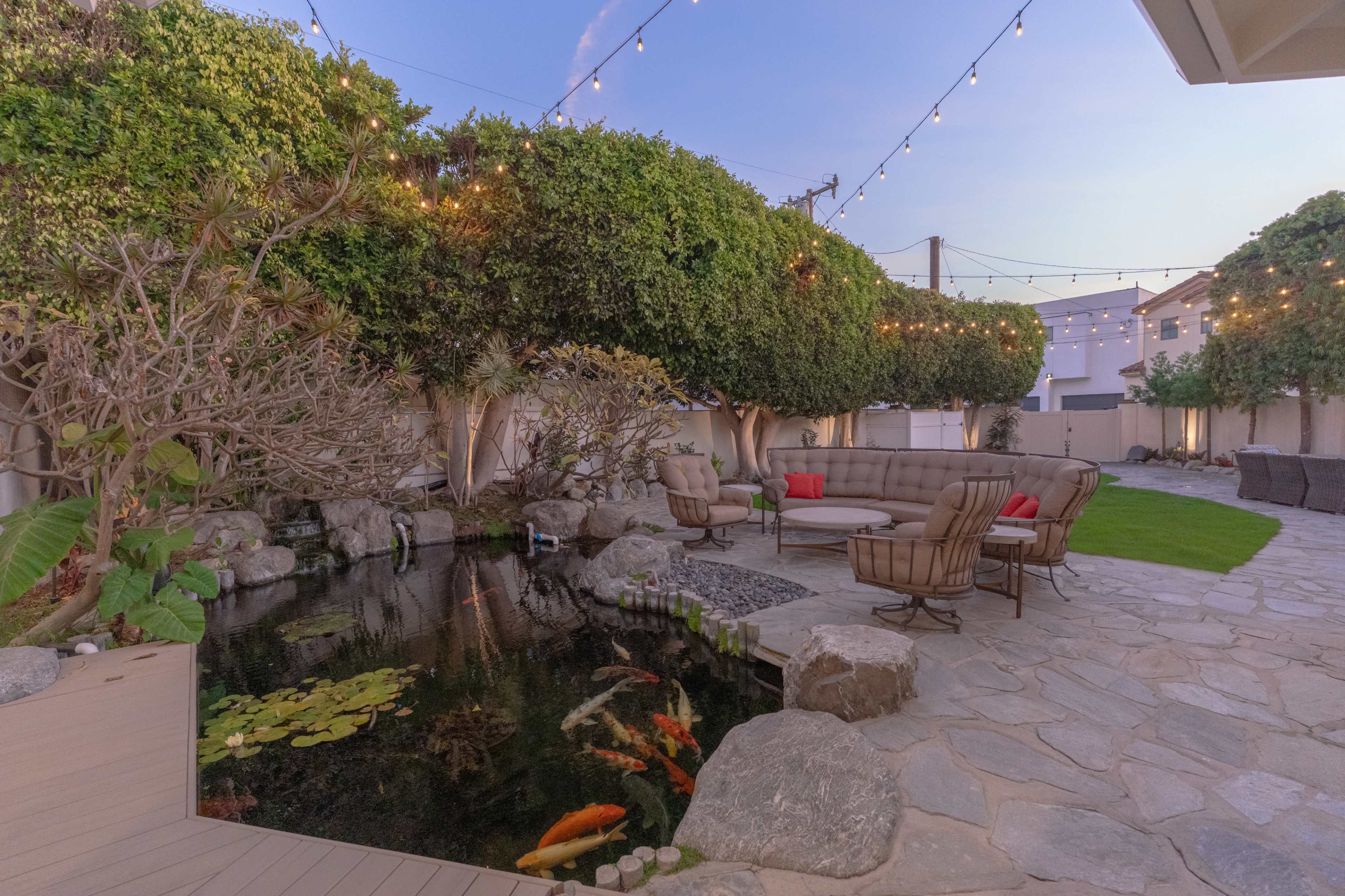 The image shows a landscaped backyard with a koi pond, rock features, seating areas, and string lights illuminating the space at dusk.