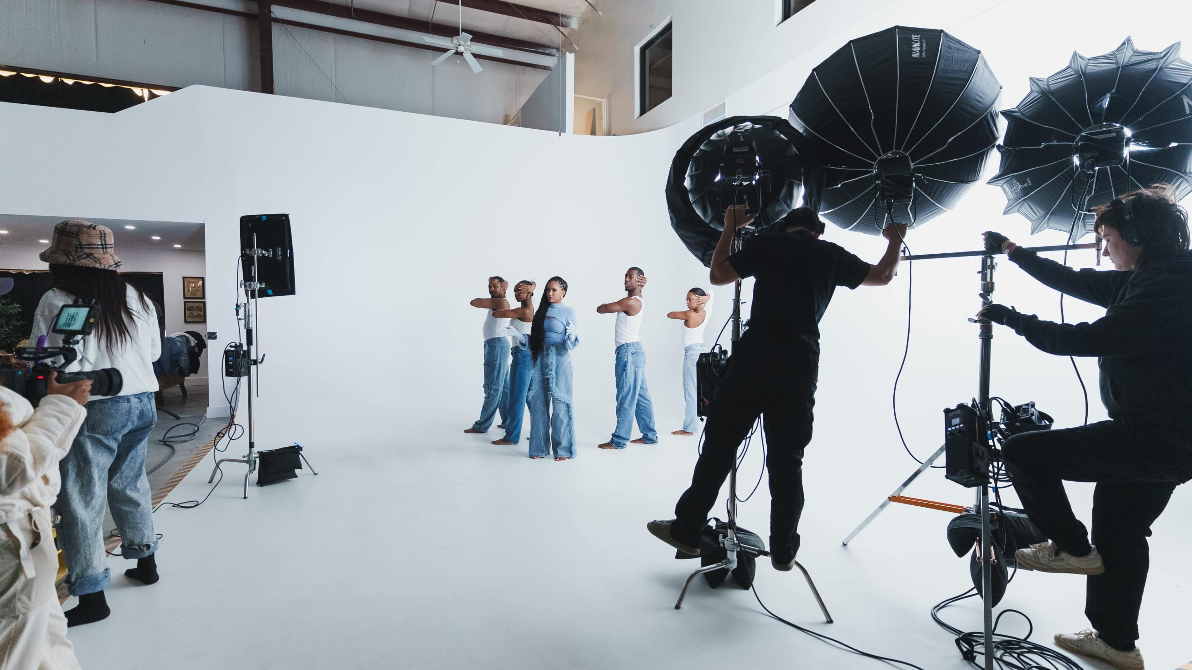 A group of models poses on a white photography set while crew members operate lighting equipment.