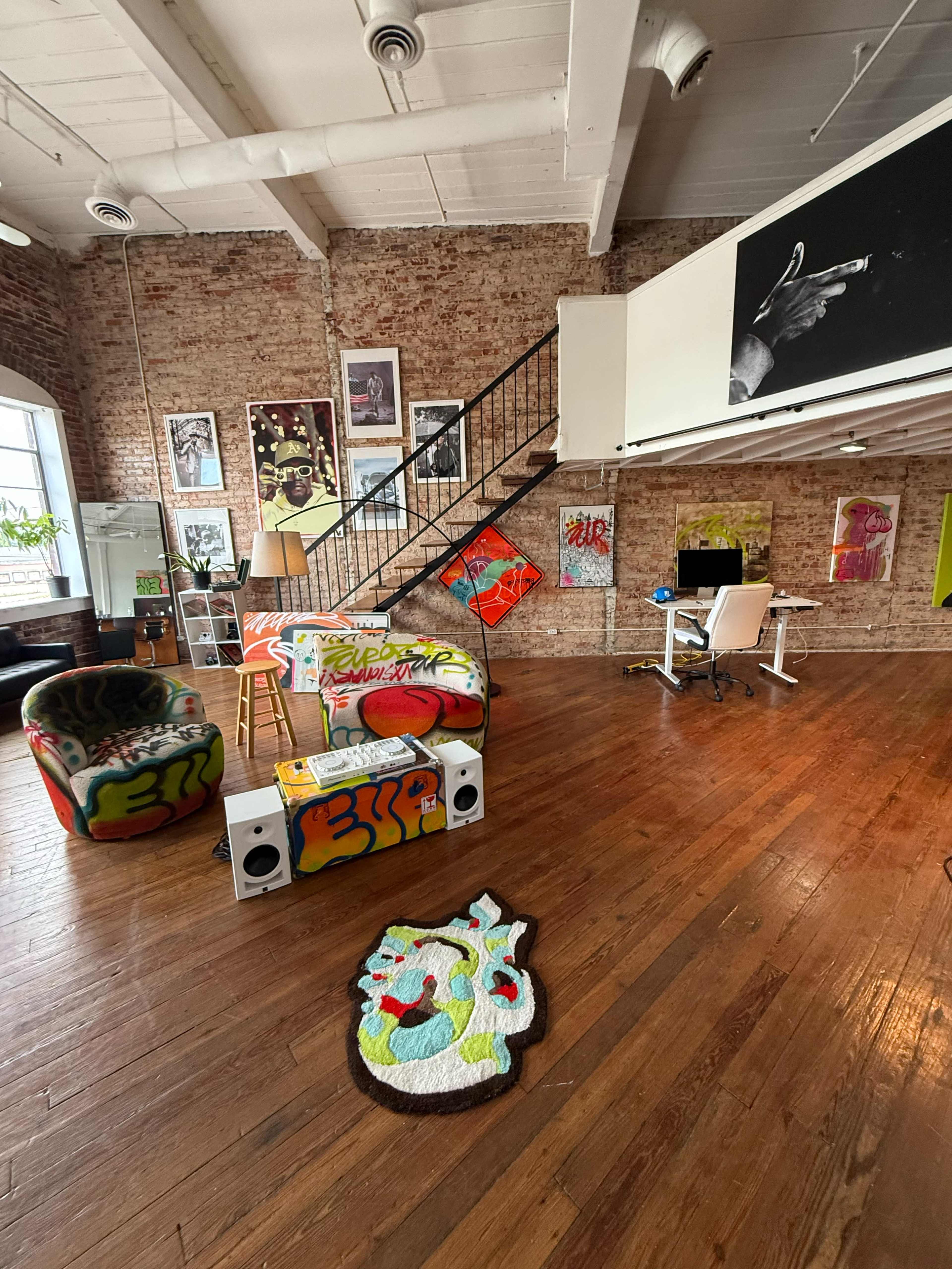 The image shows a spacious, industrial-style office with exposed brick walls, a staircase, vibrant graffiti-covered furniture, and a desk area equipped with a computer.