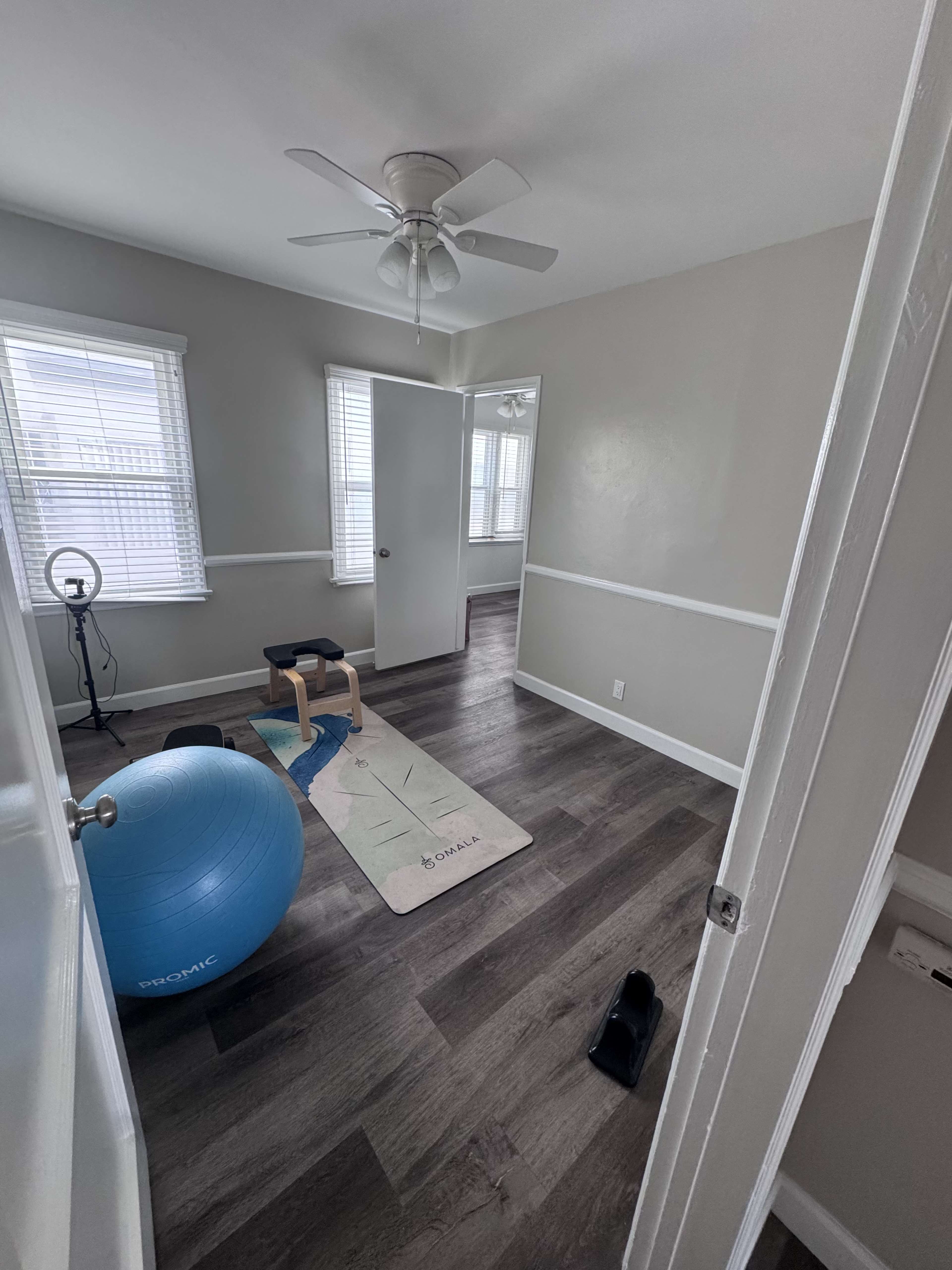 The image shows a room with a large exercise ball, a yoga mat, a wooden bench, and a pair of sandals, along with natural light coming through the windows.