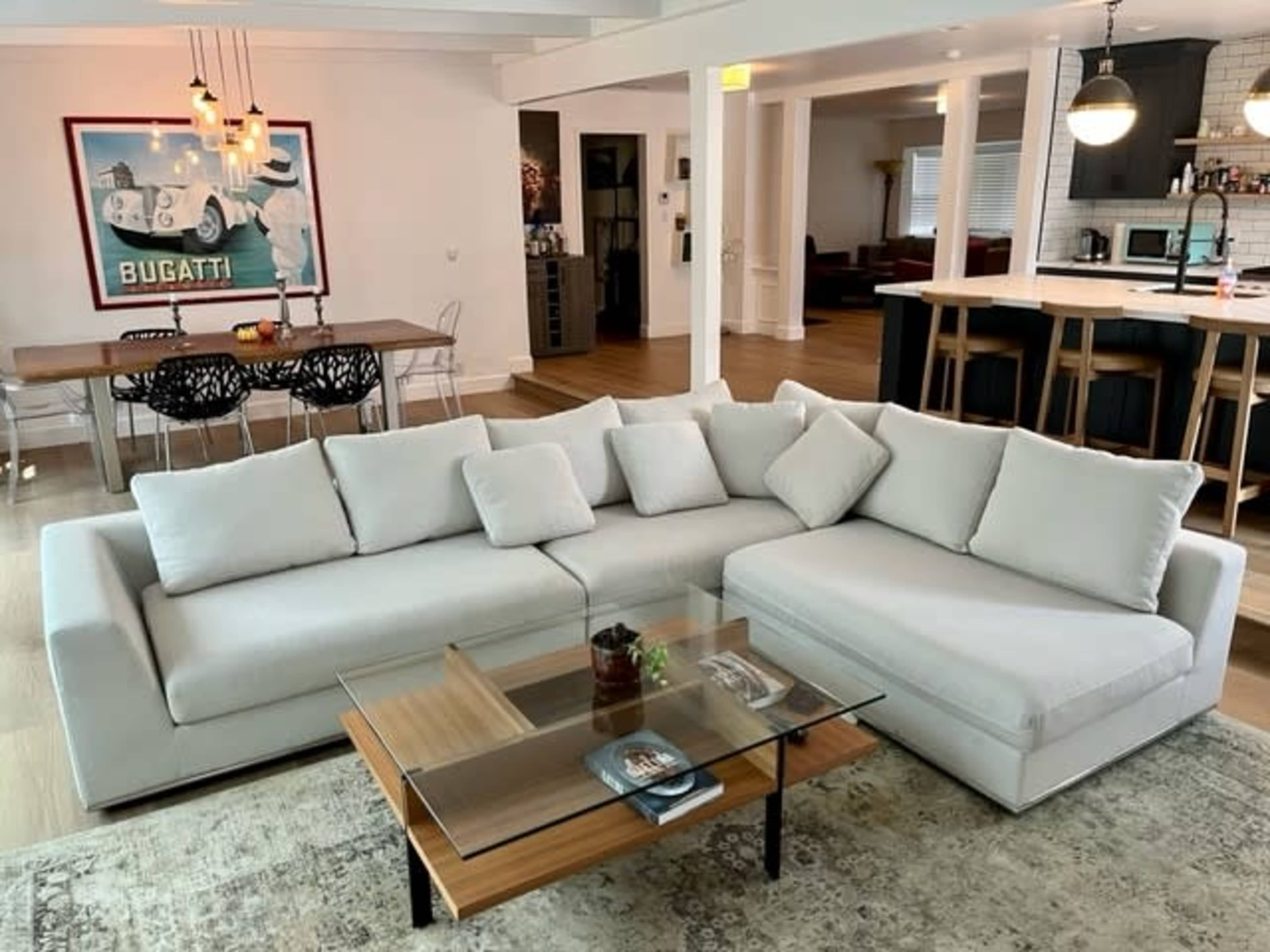 A modern living room features a light gray sectional sofa, a wooden coffee table, and a dining area with a Bugatti poster on the wall.