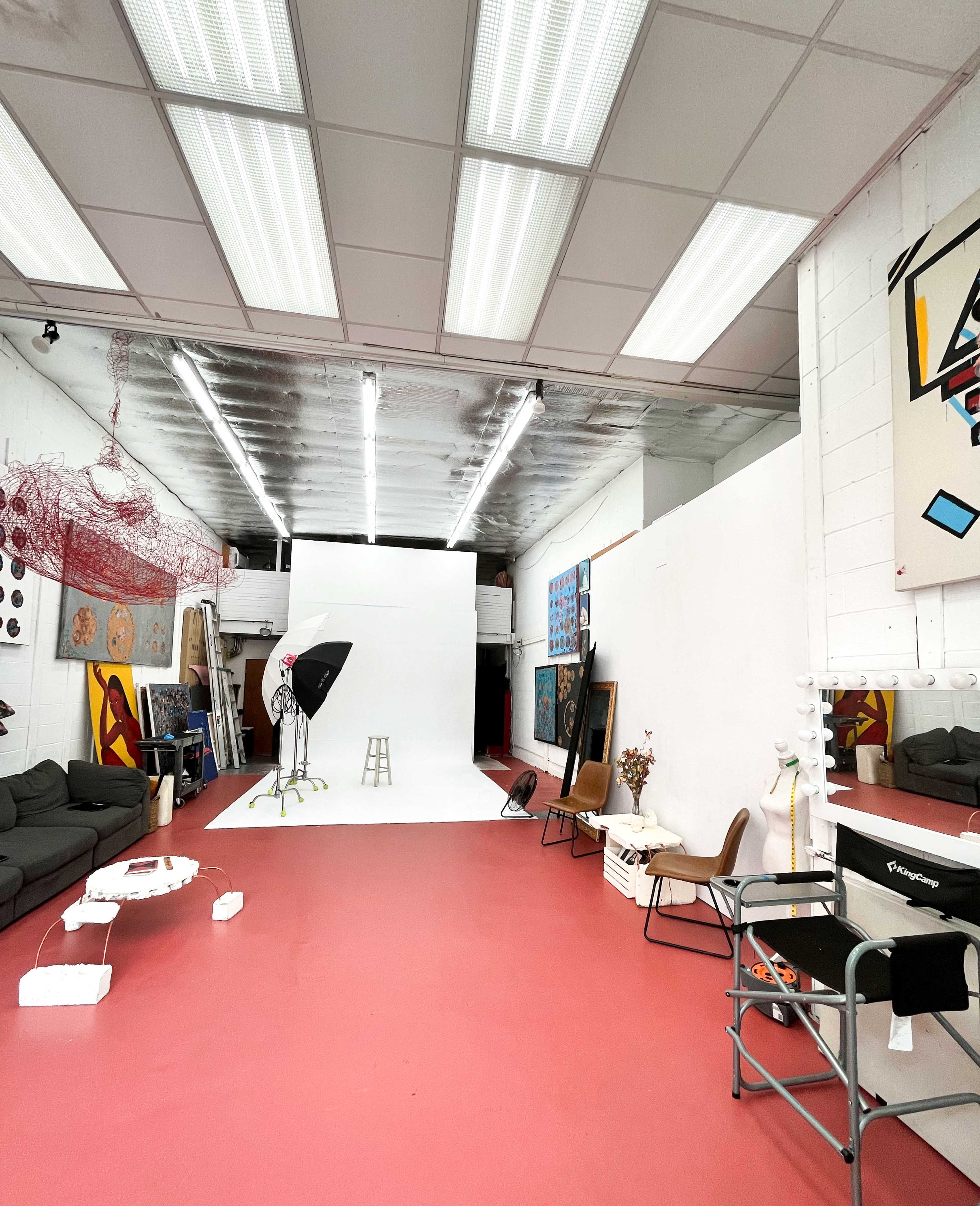 A spacious photography studio features a white backdrop, various art pieces on the walls, and seating arrangements on a red floor.