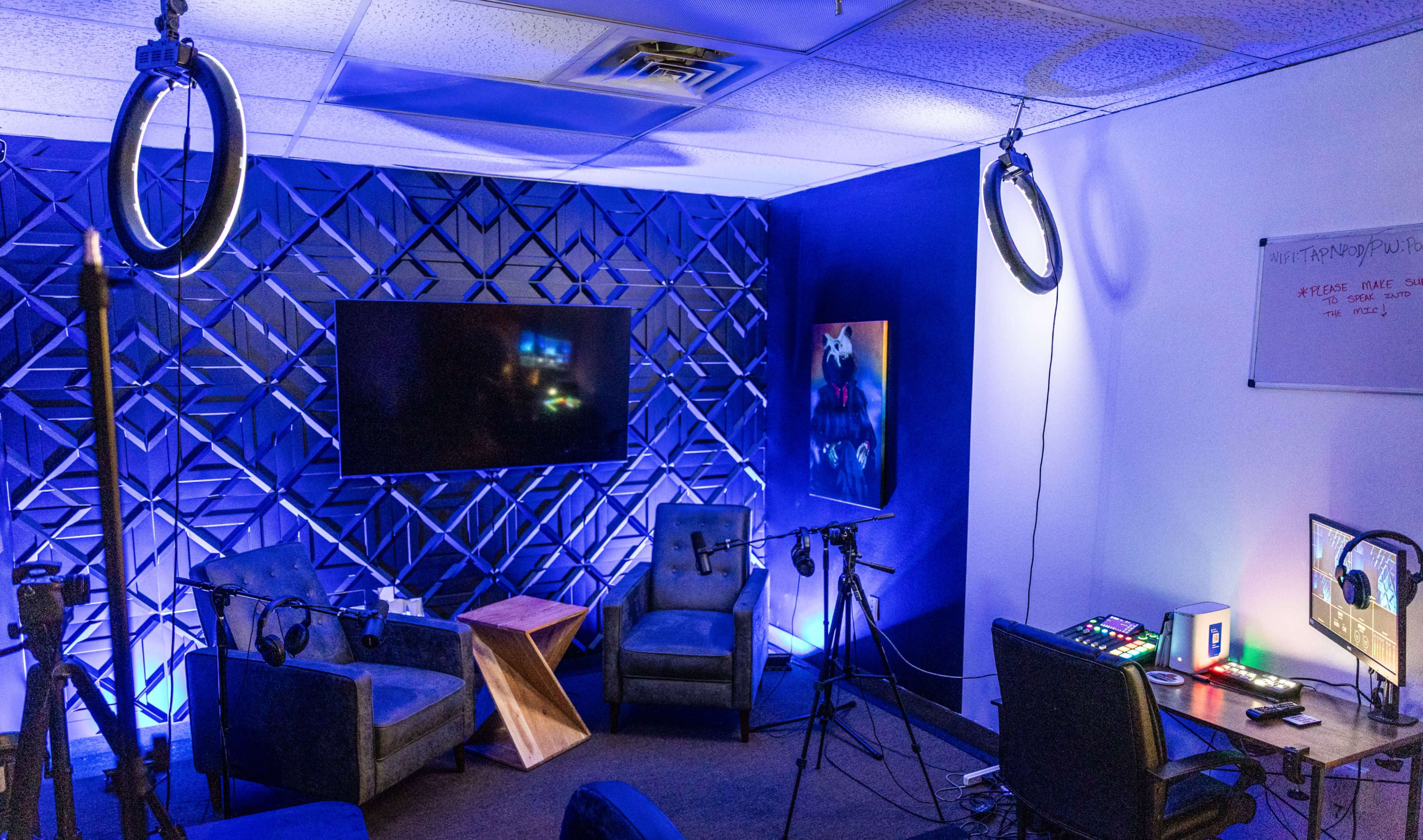 The image shows a modern recording studio setup with two chairs, a television on the wall, various microphones, and colorful lighting.