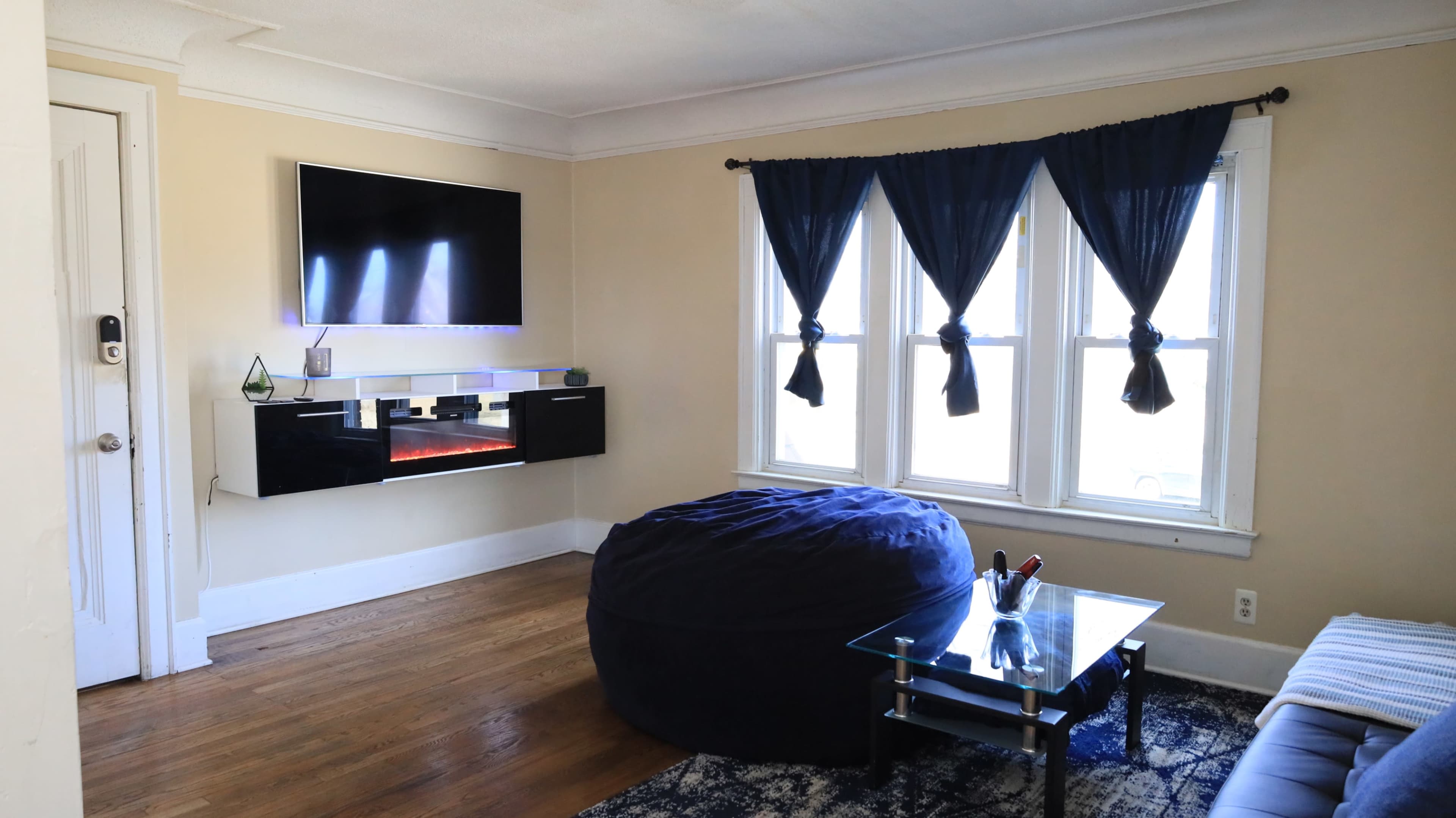 The room features a wall-mounted television, a large bean bag, and a glass coffee table, with three windows covered by dark curtains.