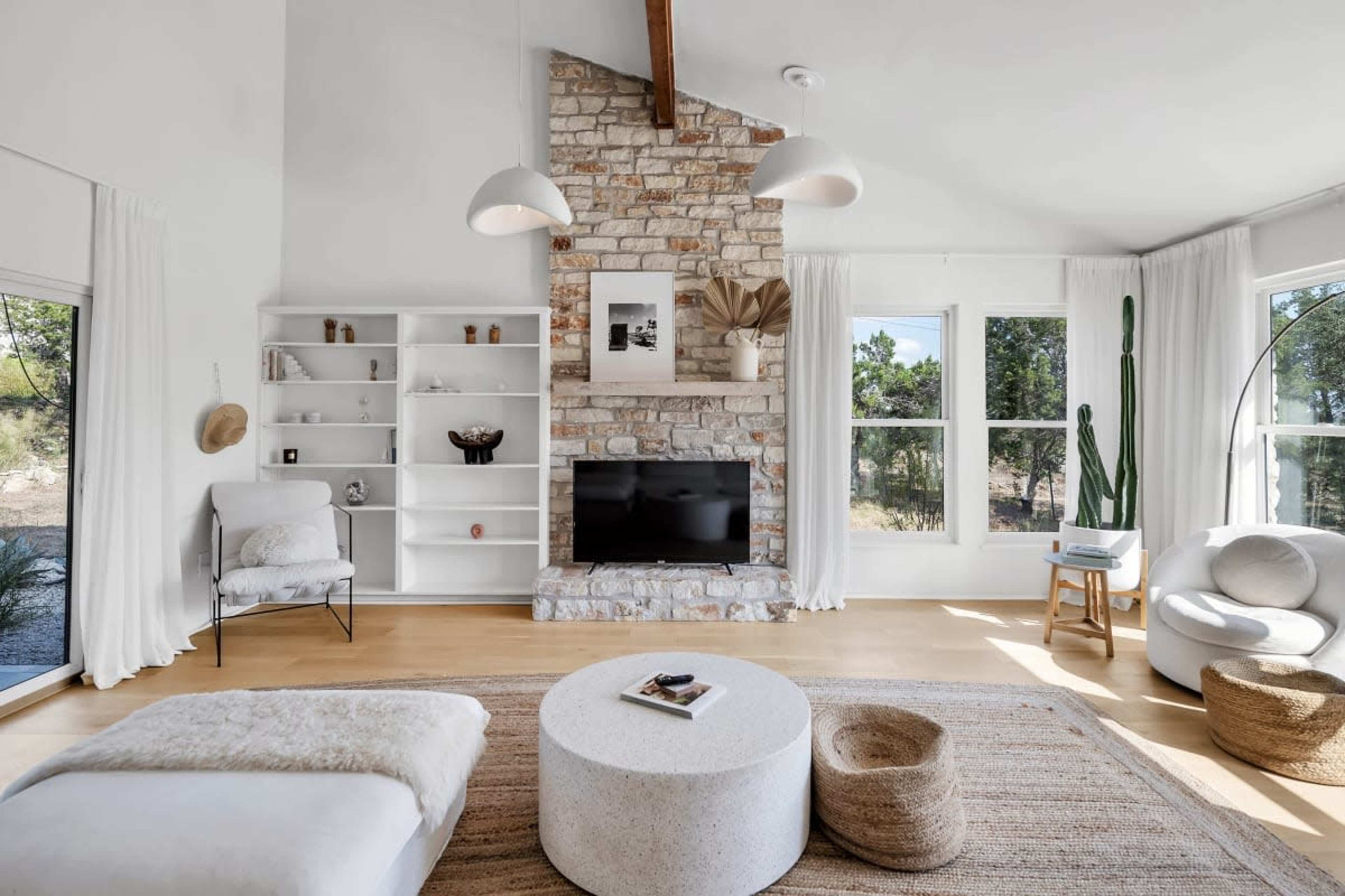 A bright living room features a stone fireplace, large windows, and minimalist furnishings, including a round coffee table and a cozy chair.