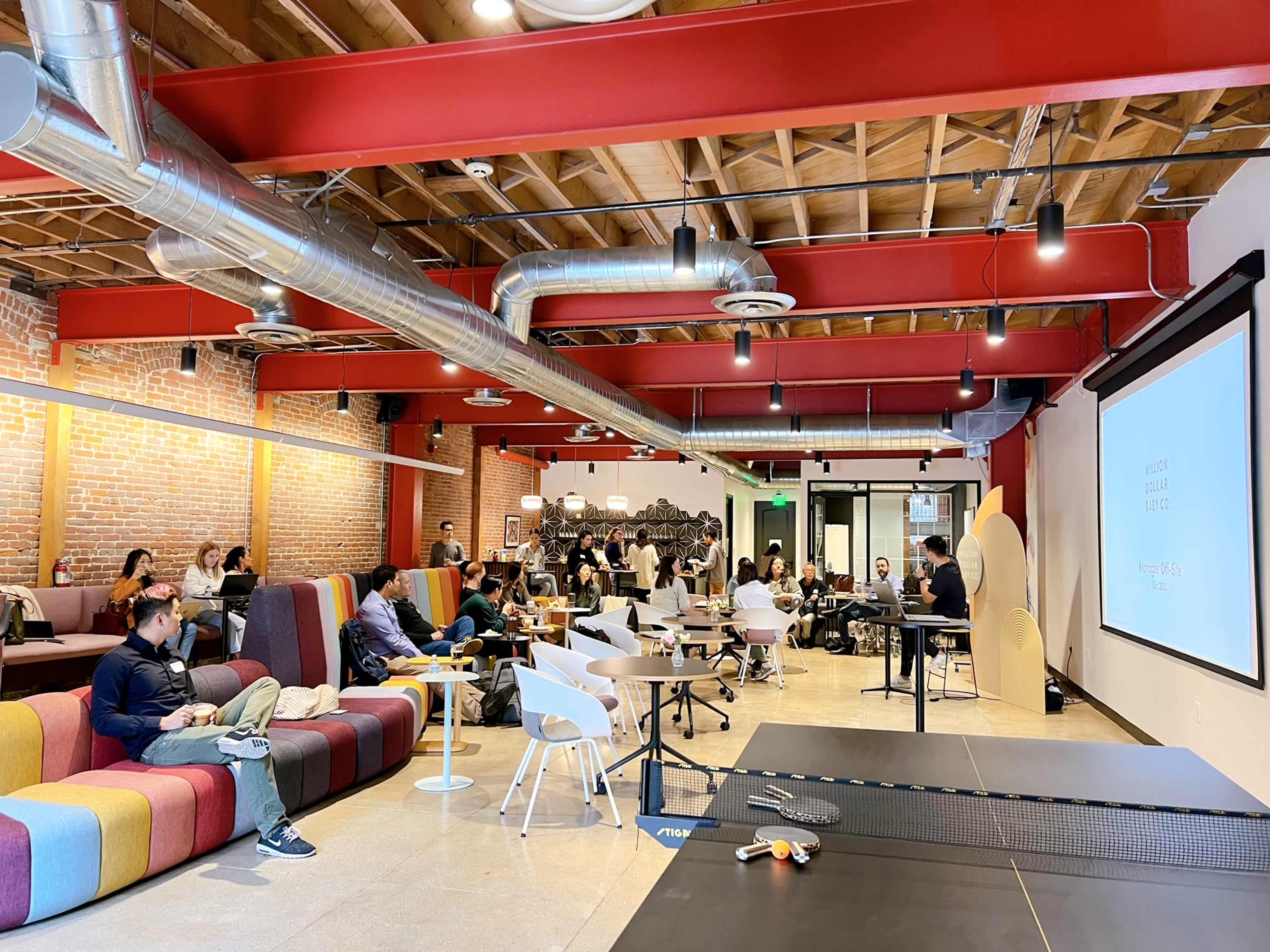 A spacious, modern co-working space filled with people seated on colorful sofas, chairs, and tables, with a projector screen displaying a presentation.
