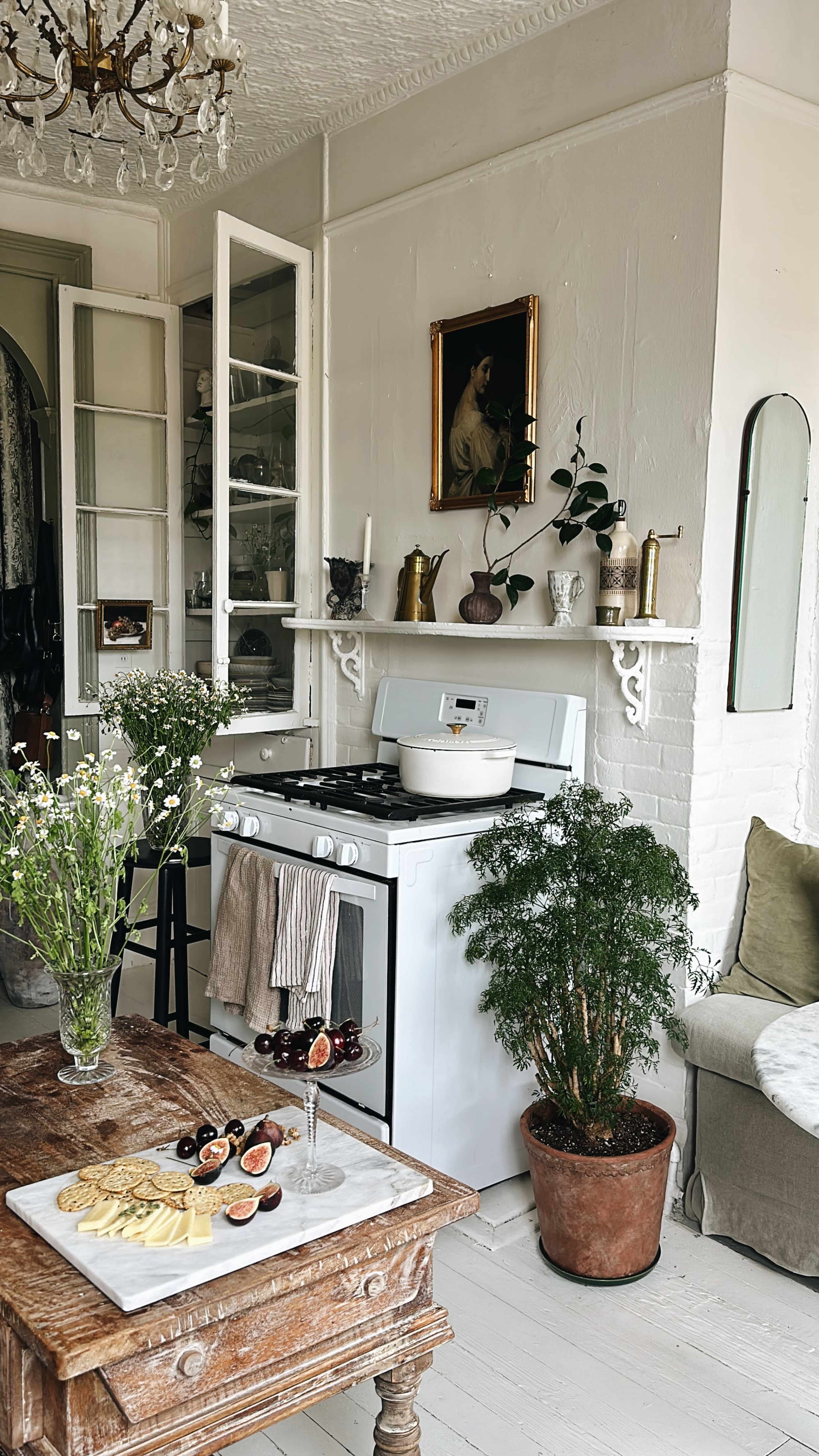 A vintage kitchen features a gas stove, white shelves with various kitchenware, and a wooden table adorned with a platter of cheese and fruits, complemented by potted plants and fresh flowers.
