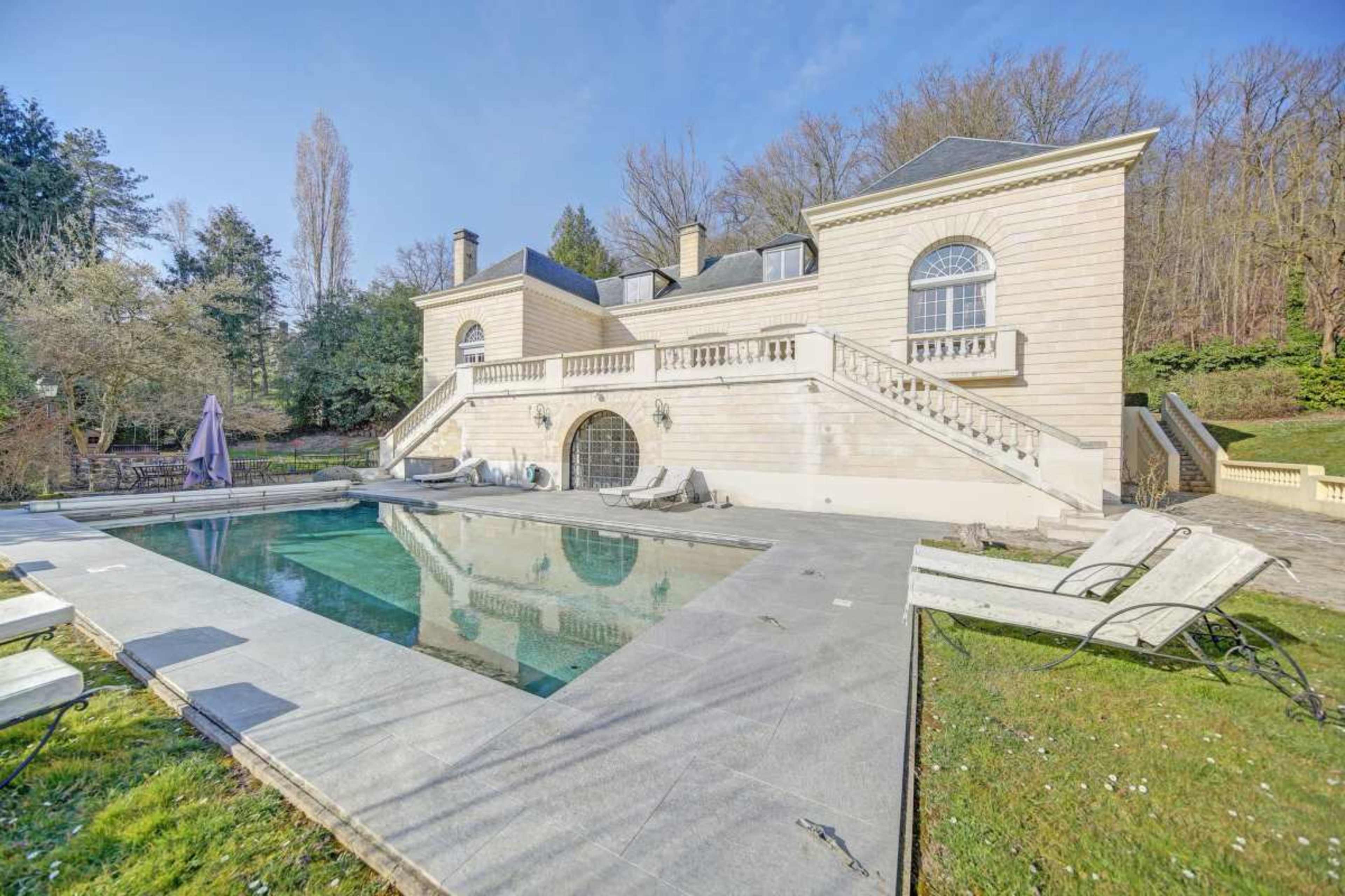 The image shows a large, elegant stone house with a terrace overlooking a rectangular swimming pool, surrounded by green grass and trees.