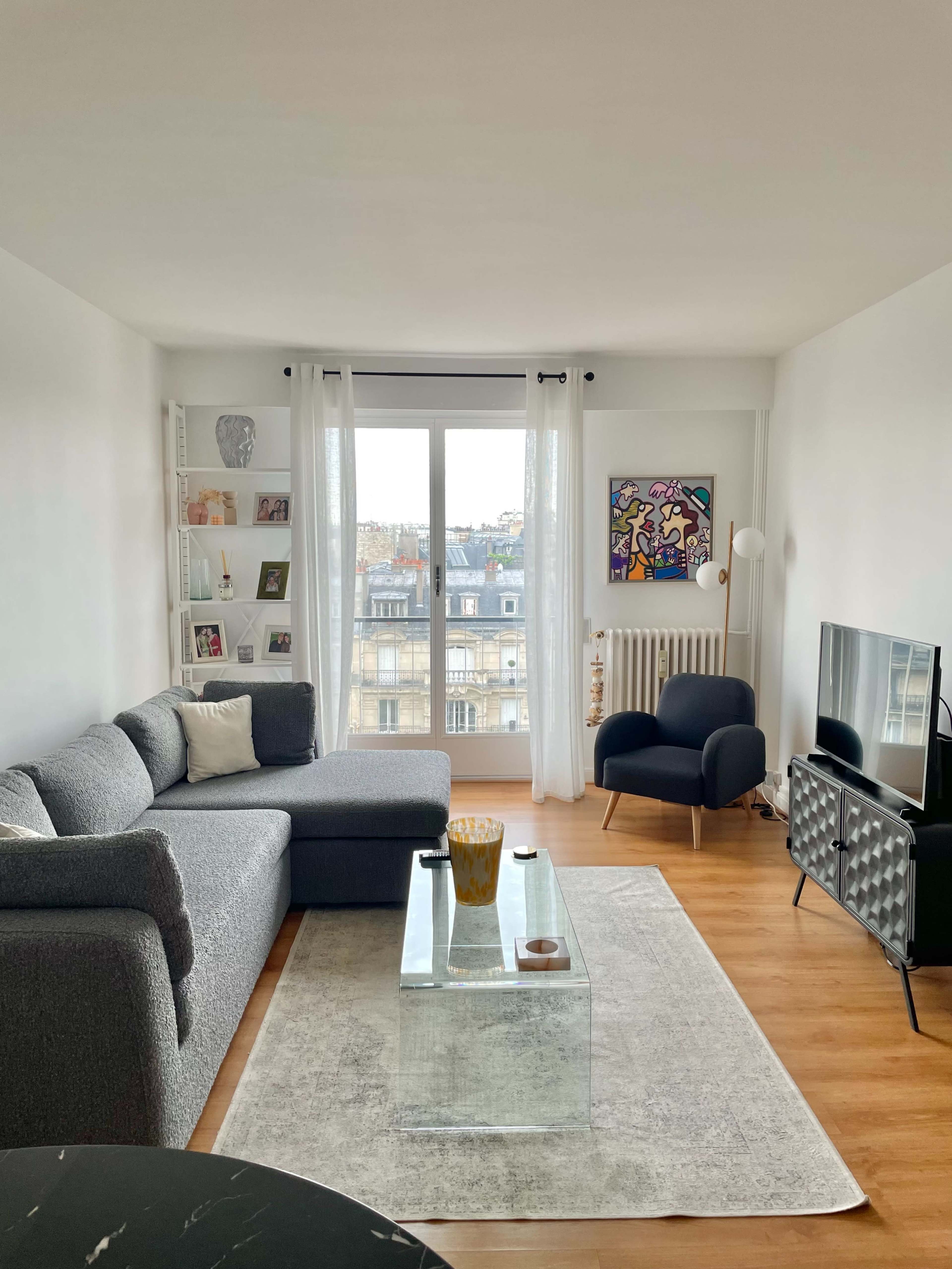 A modern living room features a gray sectional sofa, a glass coffee table, and a black armchair, with large windows providing a view of city rooftops.