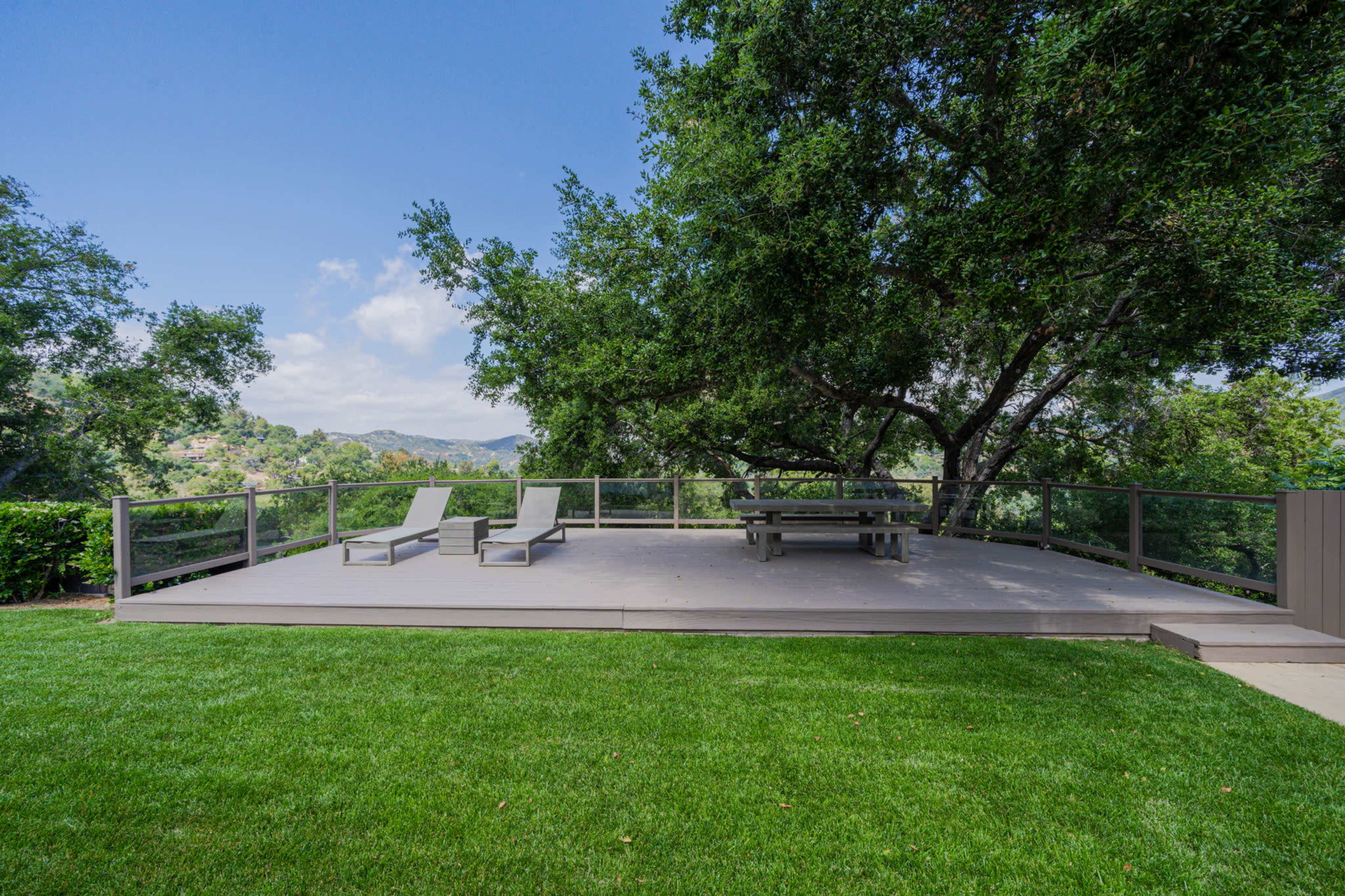 A wooden deck with lounge chairs and a picnic table overlooks a green lawn and surrounding trees.