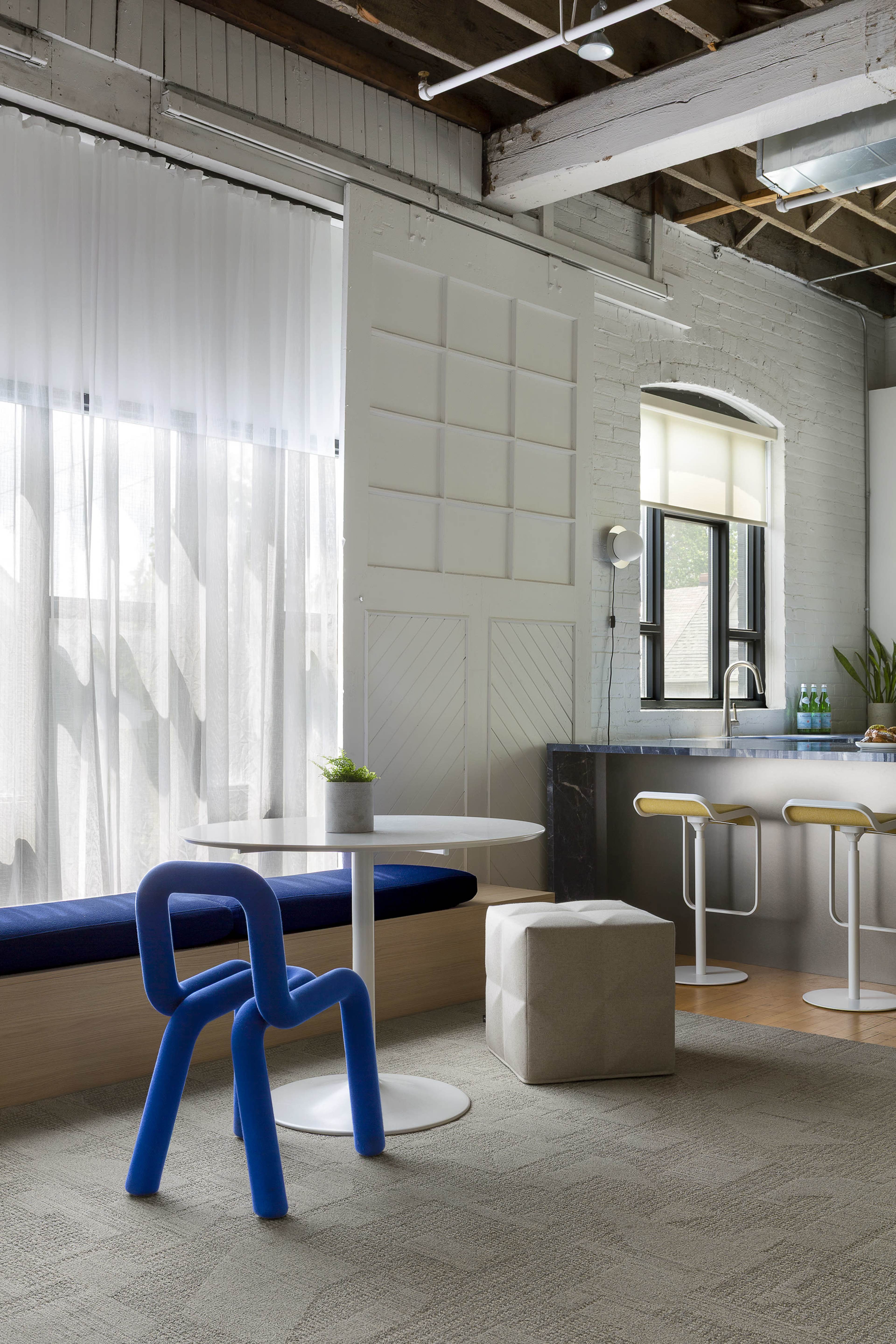 The image shows a modern interior space with a blue chair, a round white table, a gray ottoman, and a kitchen area featuring bar stools and large windows with sheer curtains.