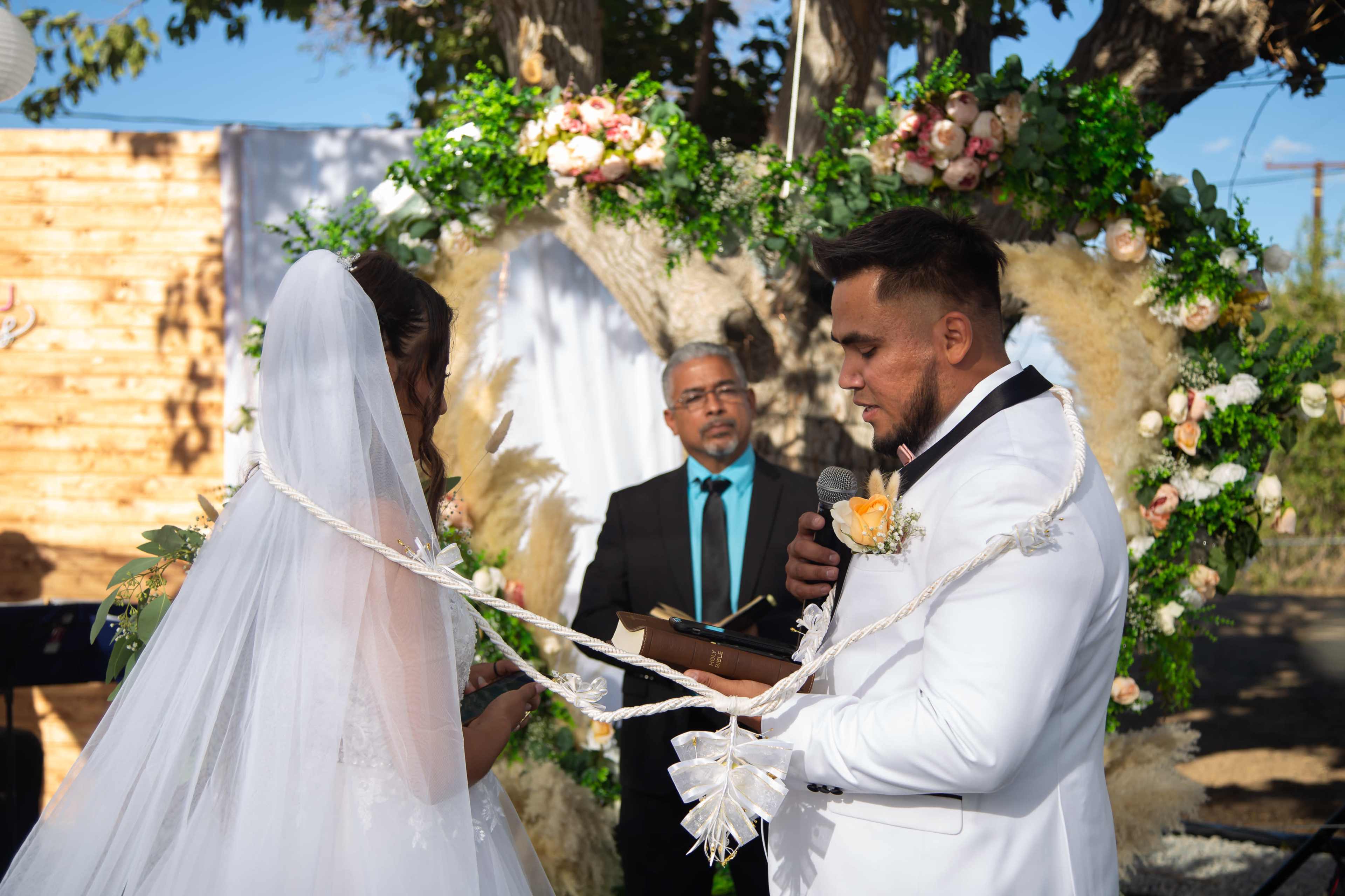 A couple exchanges vows during a wedding ceremony, while a officiant stands in front of them and a floral arch frames the scene.