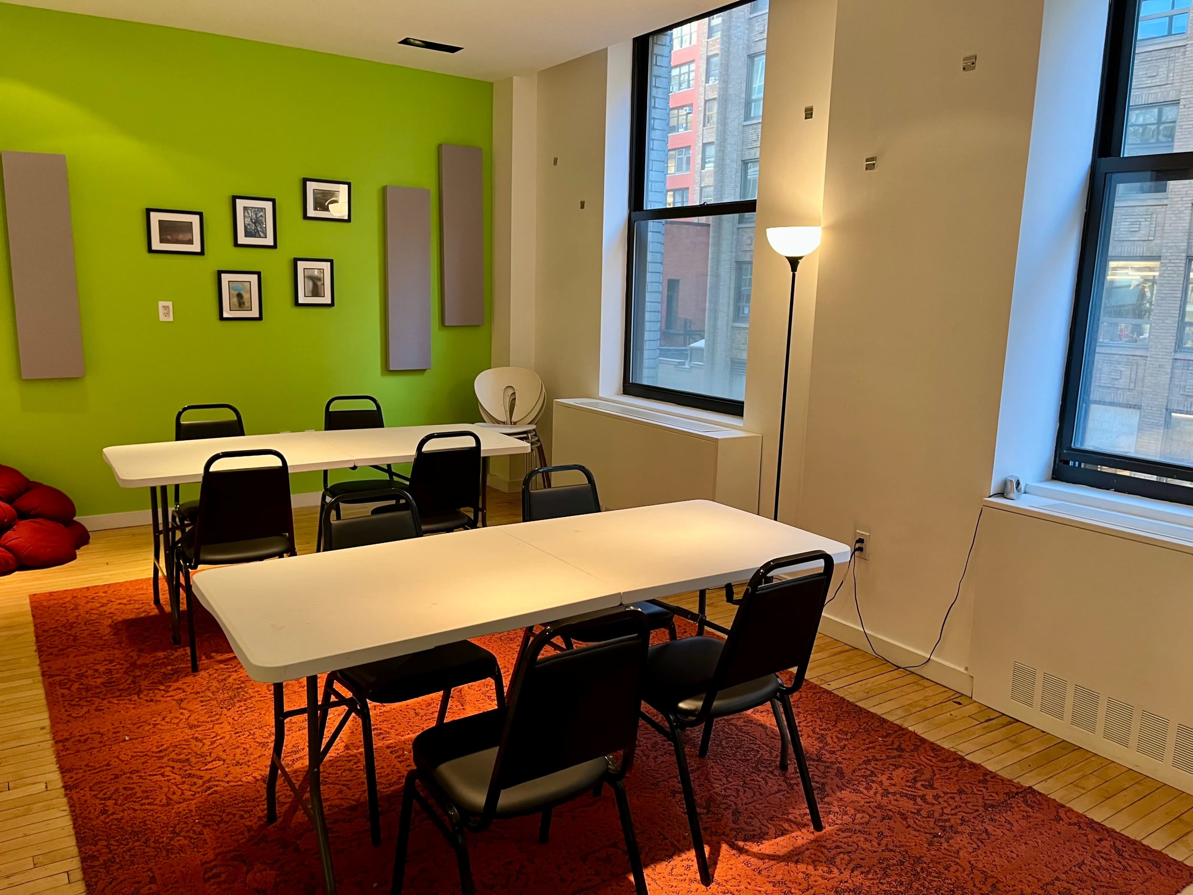 The room features several folding tables and chairs arranged on a red carpet, with green walls adorned with framed pictures and large windows letting in natural light.
