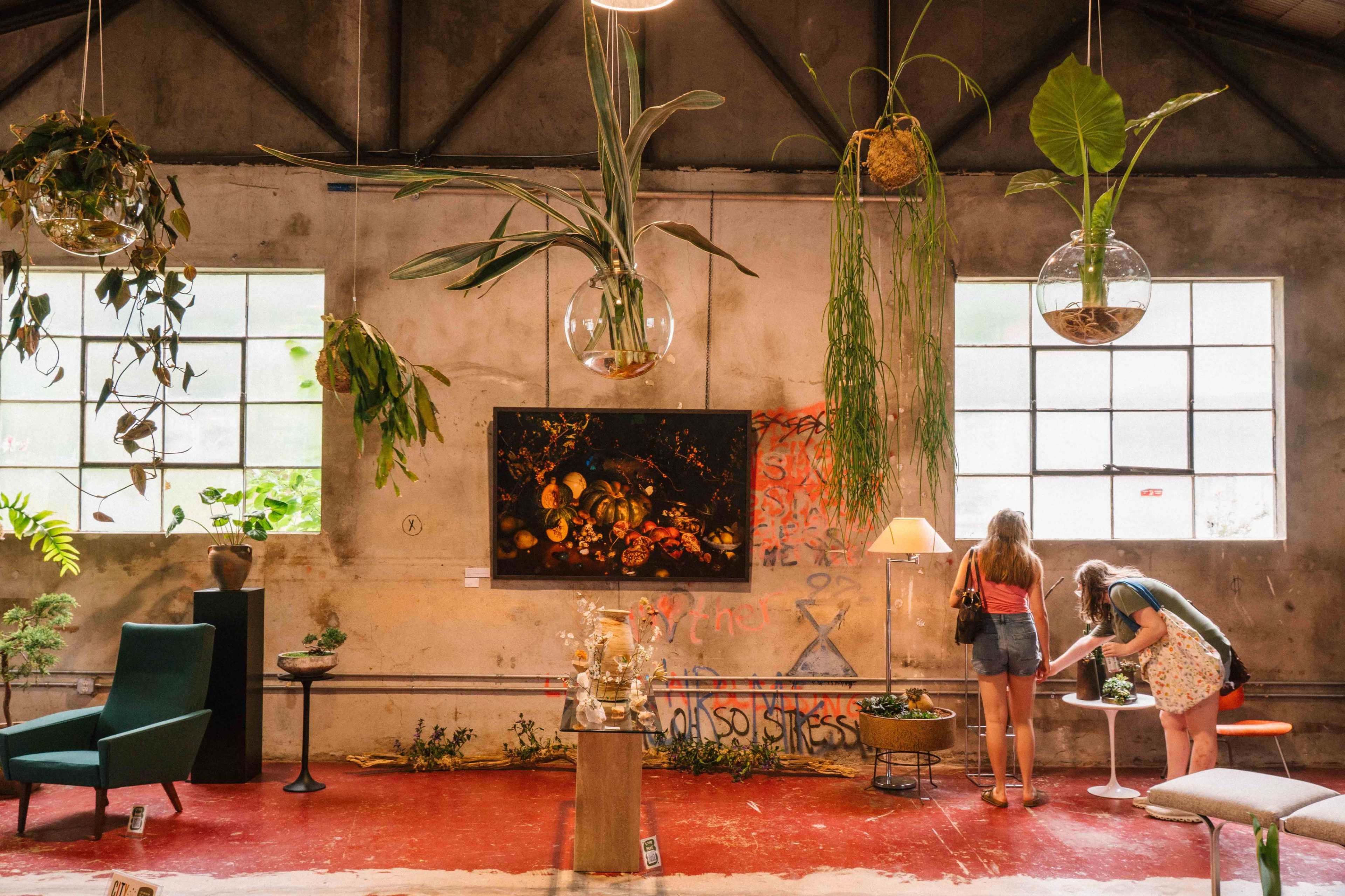 A spacious interior features a diverse arrangement of houseplants, wall art, and furniture, with two people exploring the décor.