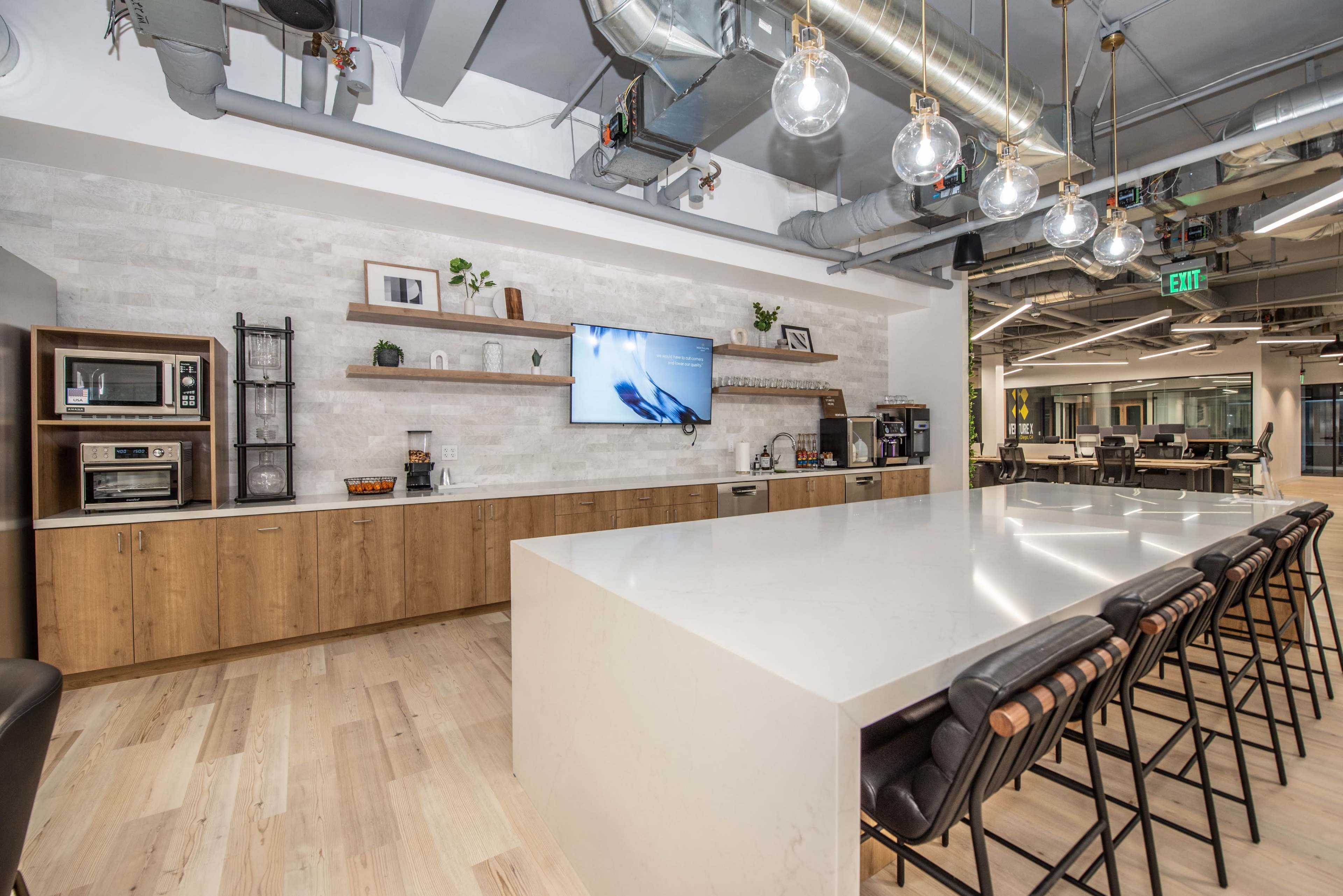 A modern office kitchen area featuring a large white countertop, wooden cabinetry, a microwave, and a television mounted on the wall.