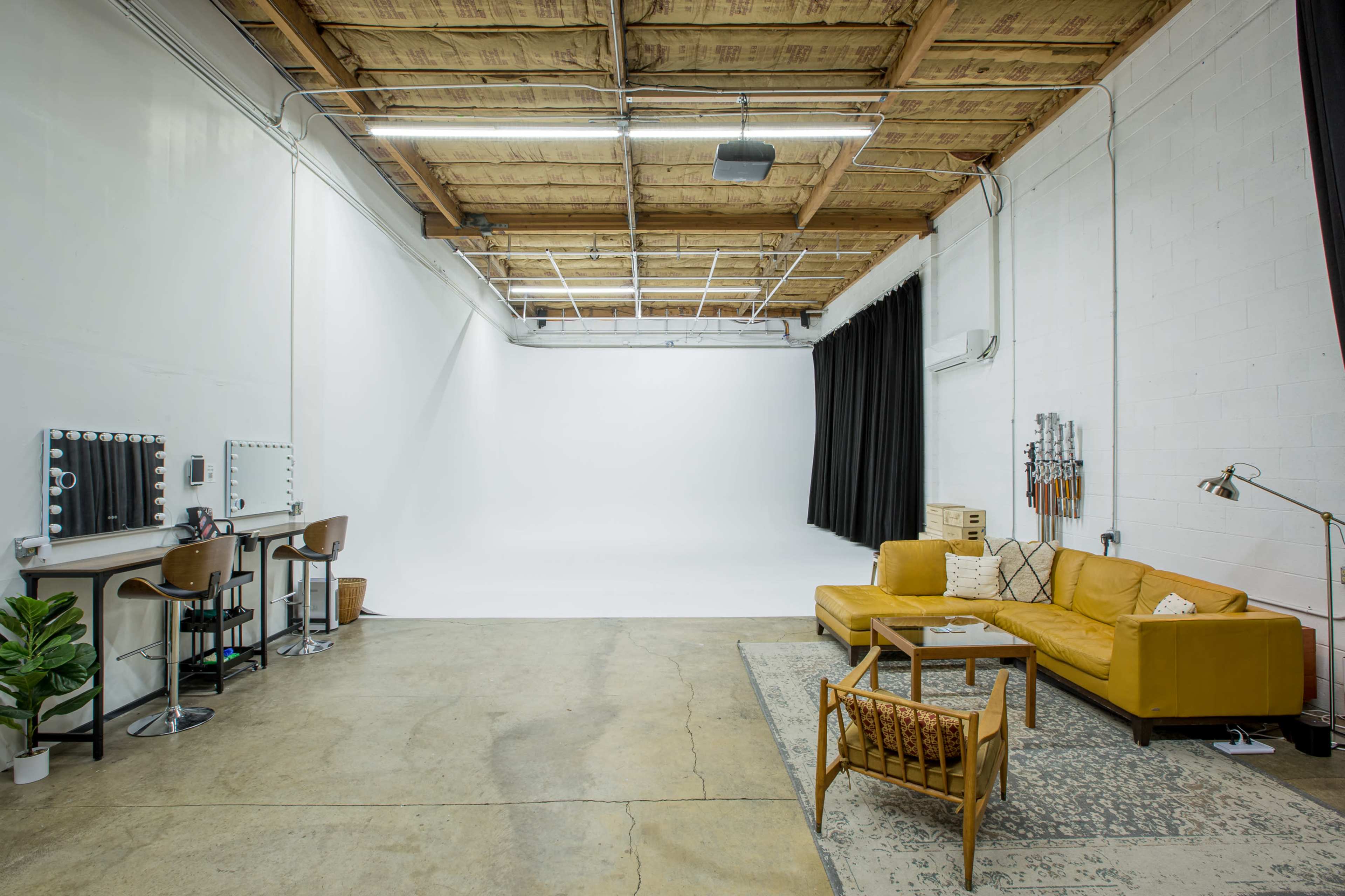 A spacious studio features a yellow sectional sofa, a wooden chair, two makeup stations with mirrors, and a backdrop of white and black curtains.