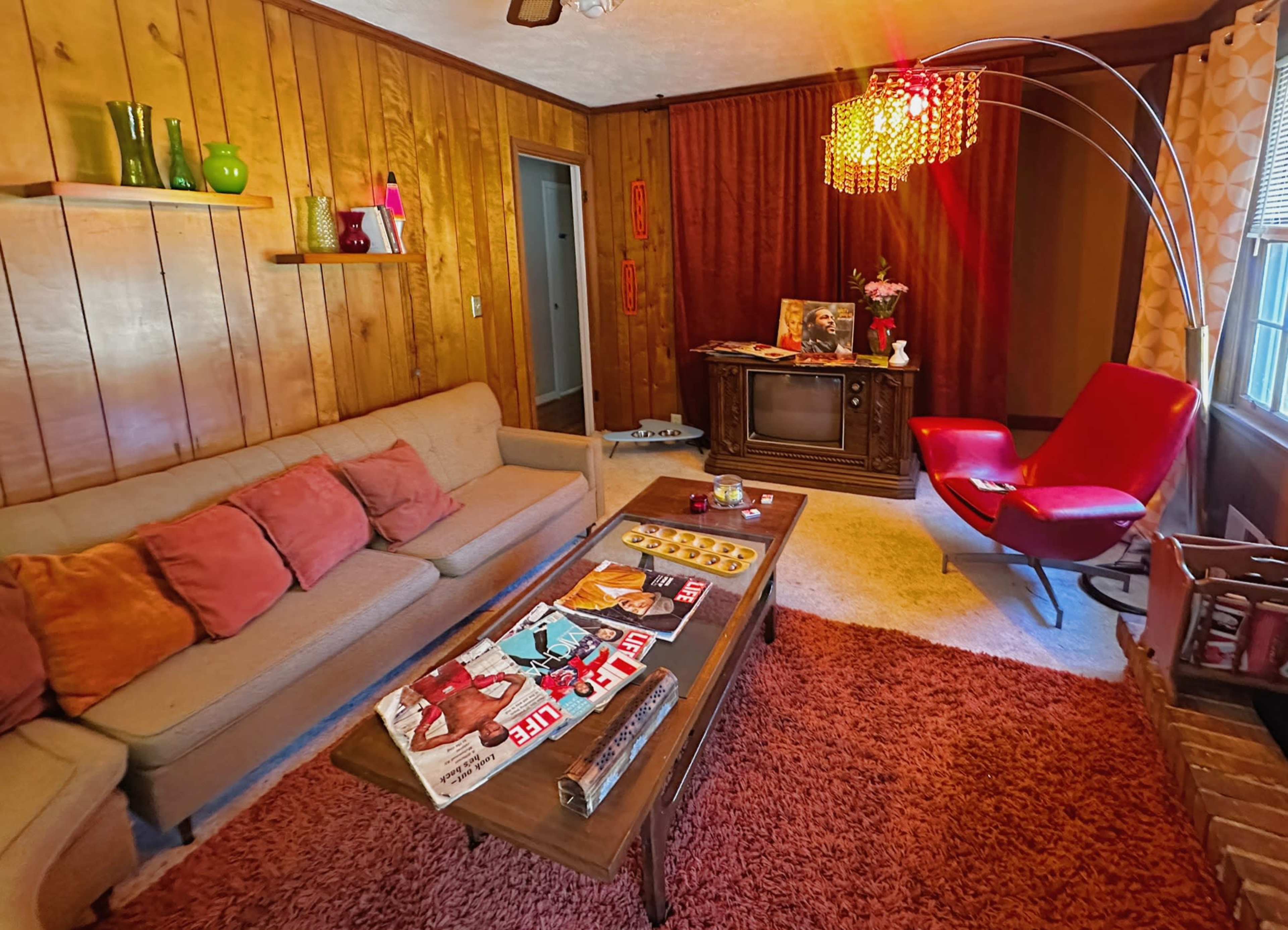 The image shows a retro living room with wooden walls, a beige sofa adorned with colorful pillows, a red armchair, a coffee table covered with magazines, and a vintage television set.