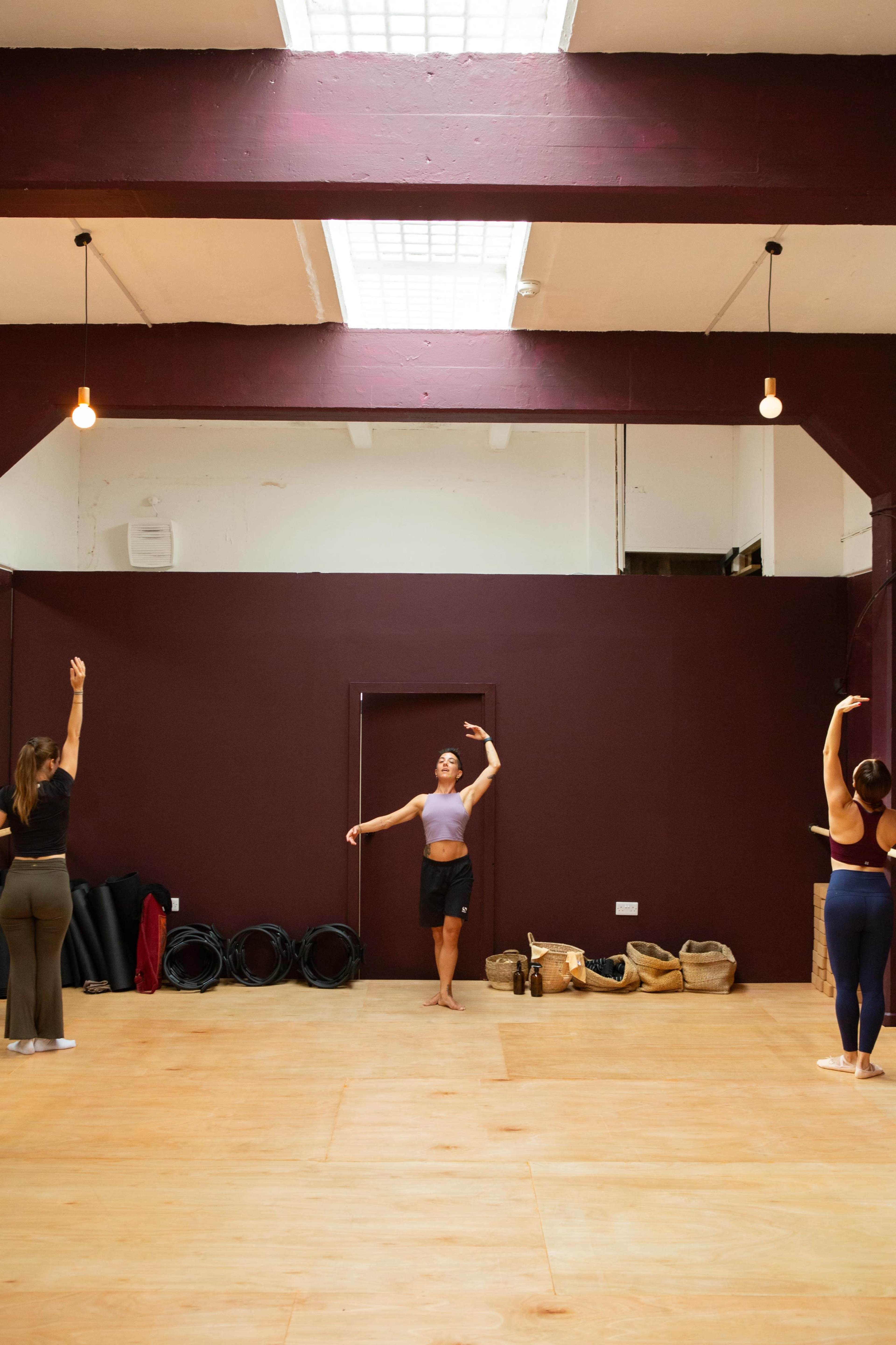 A group of four individuals is practicing dance in a spacious studio with wooden floors and maroon walls.