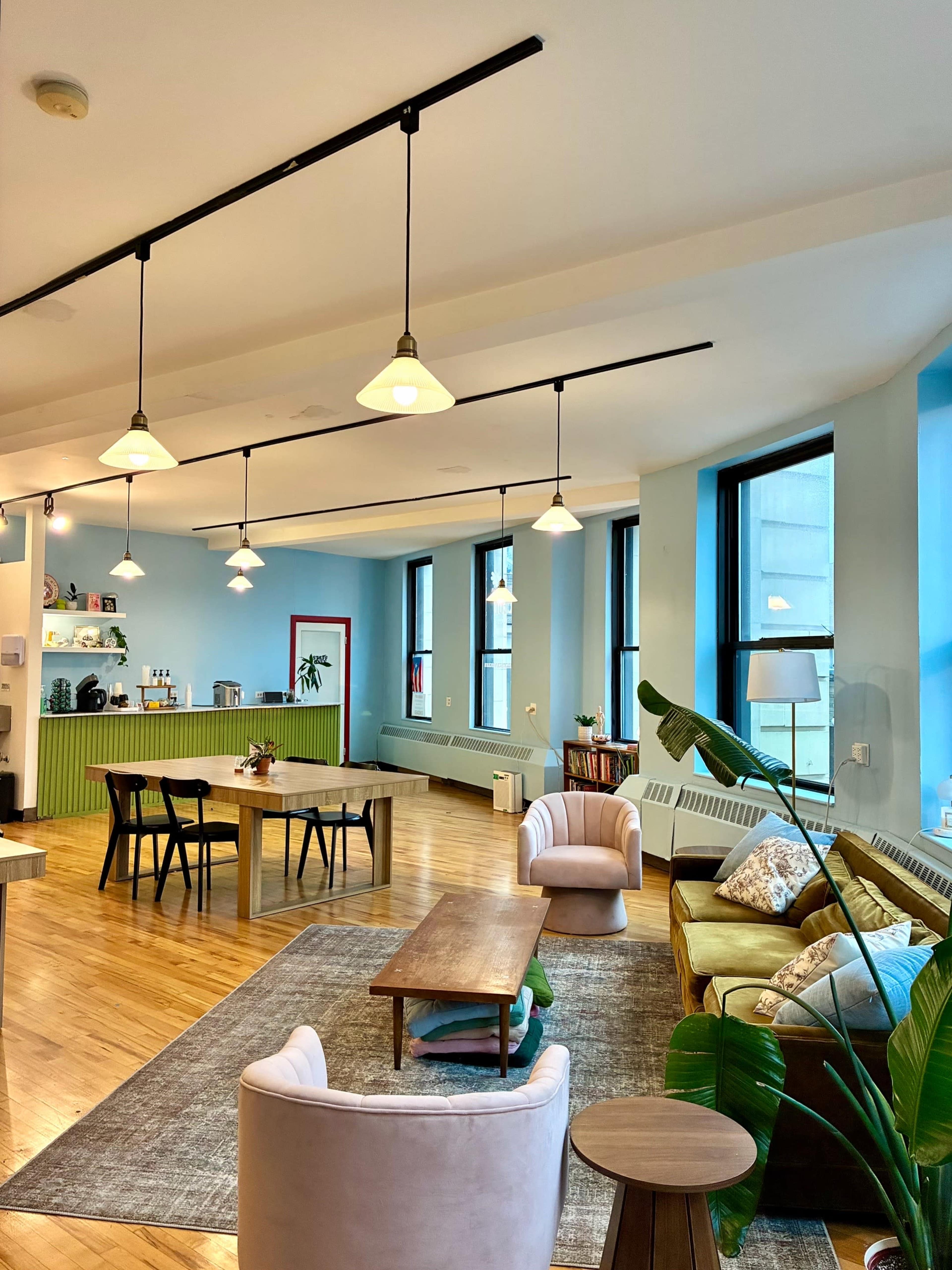 The image shows a modern office space with wooden floors, multiple windows, a green kitchen area, and various seating arrangements, including chairs and a coffee table.