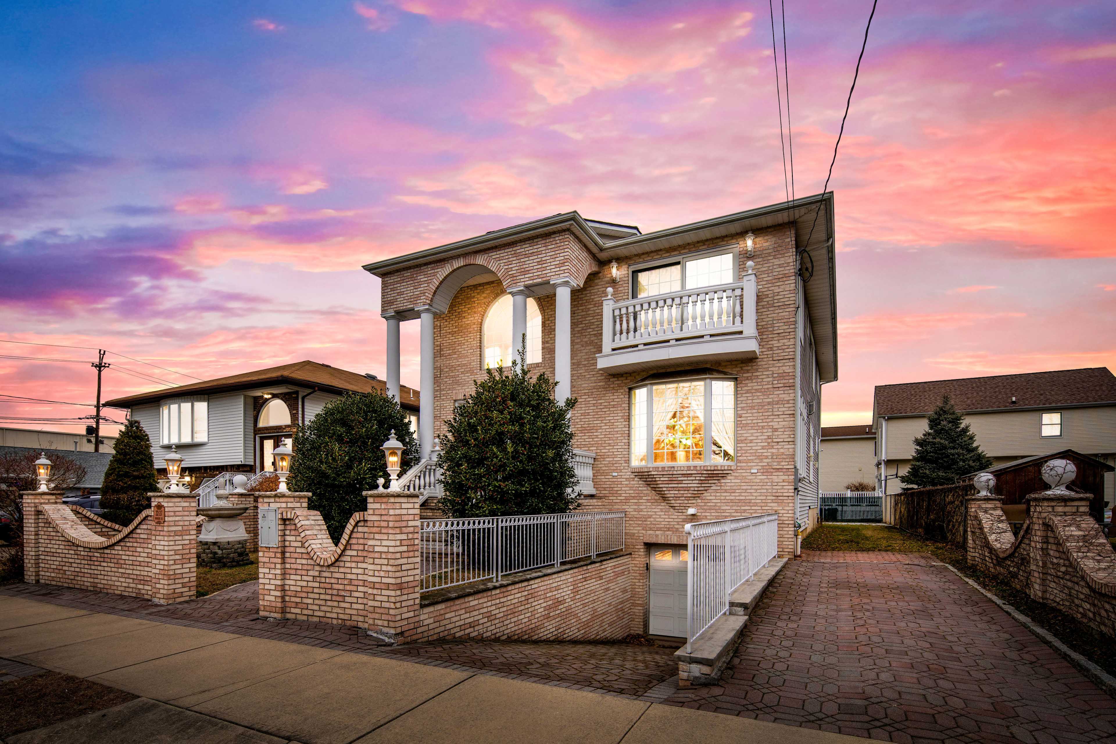 A two-story brick house with a decorative front entrance and landscaped yard is illuminated by a colorful sunset.