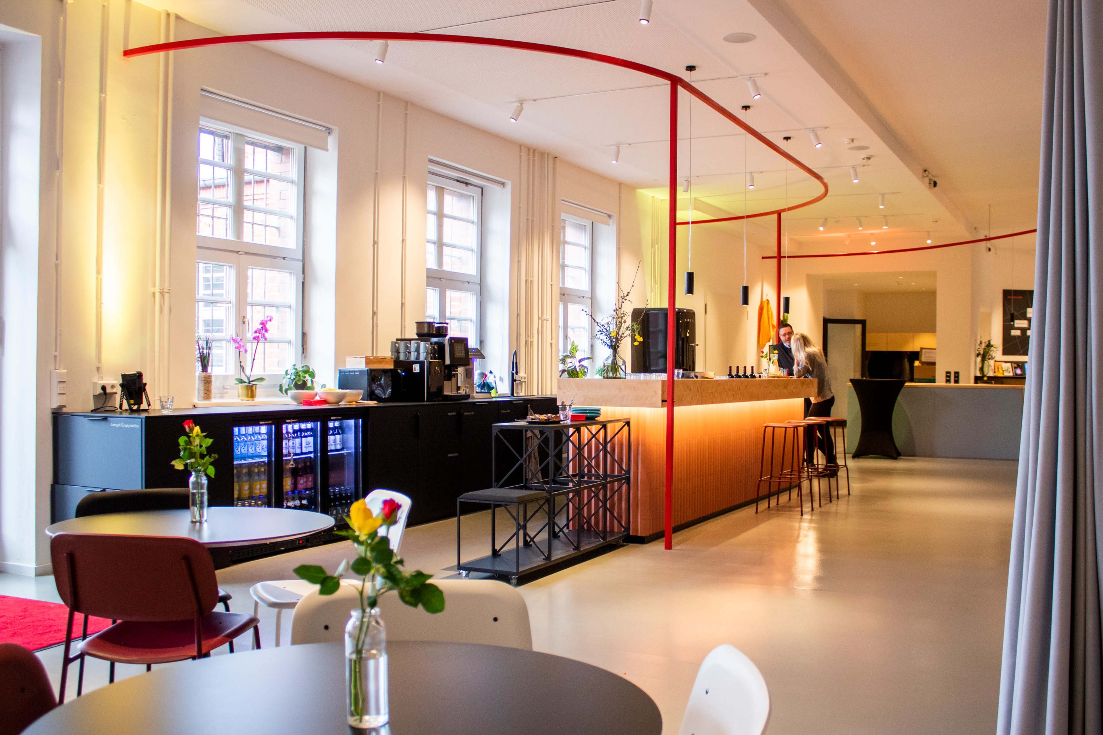 A modern café features a sleek counter with a barista, colorful shelves, and minimalist furniture arranged around tables.