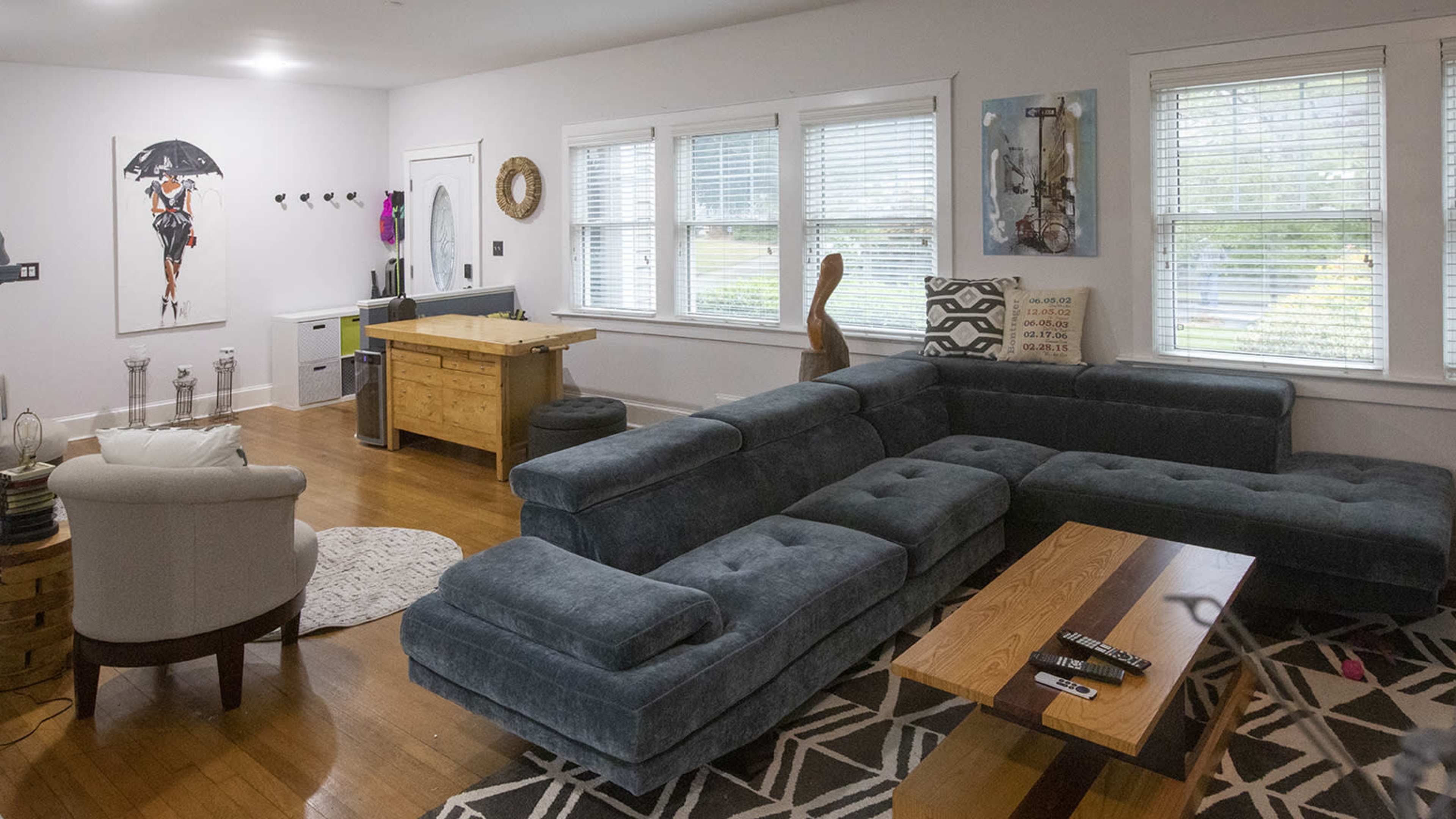 The living room features a large sectional sofa, a wooden coffee table, and a dining area with a small table, surrounded by windows with blinds.