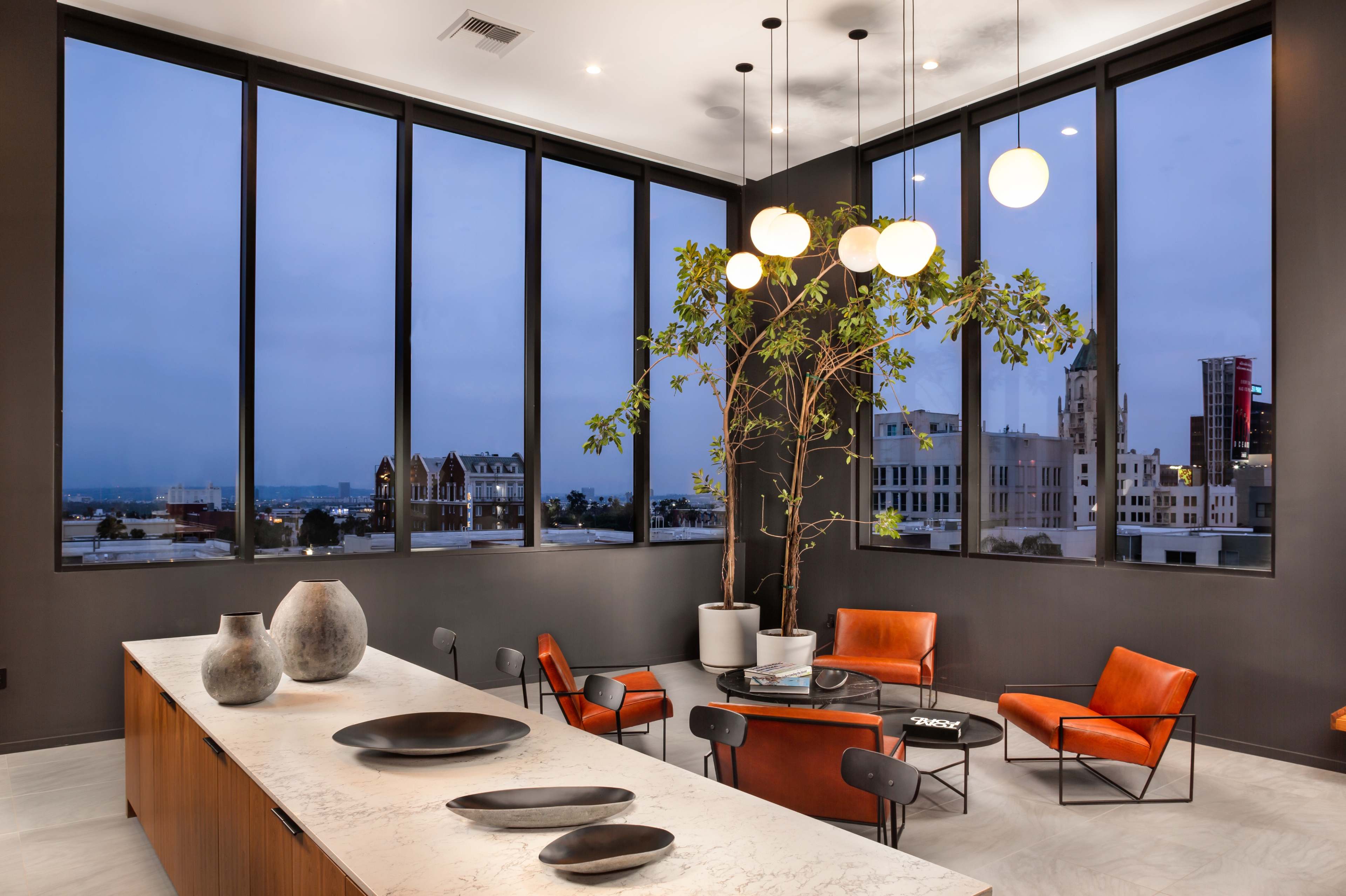 A modern lounge with a marble countertop, leather chairs, and large windows overlooking a cityscape at dusk.