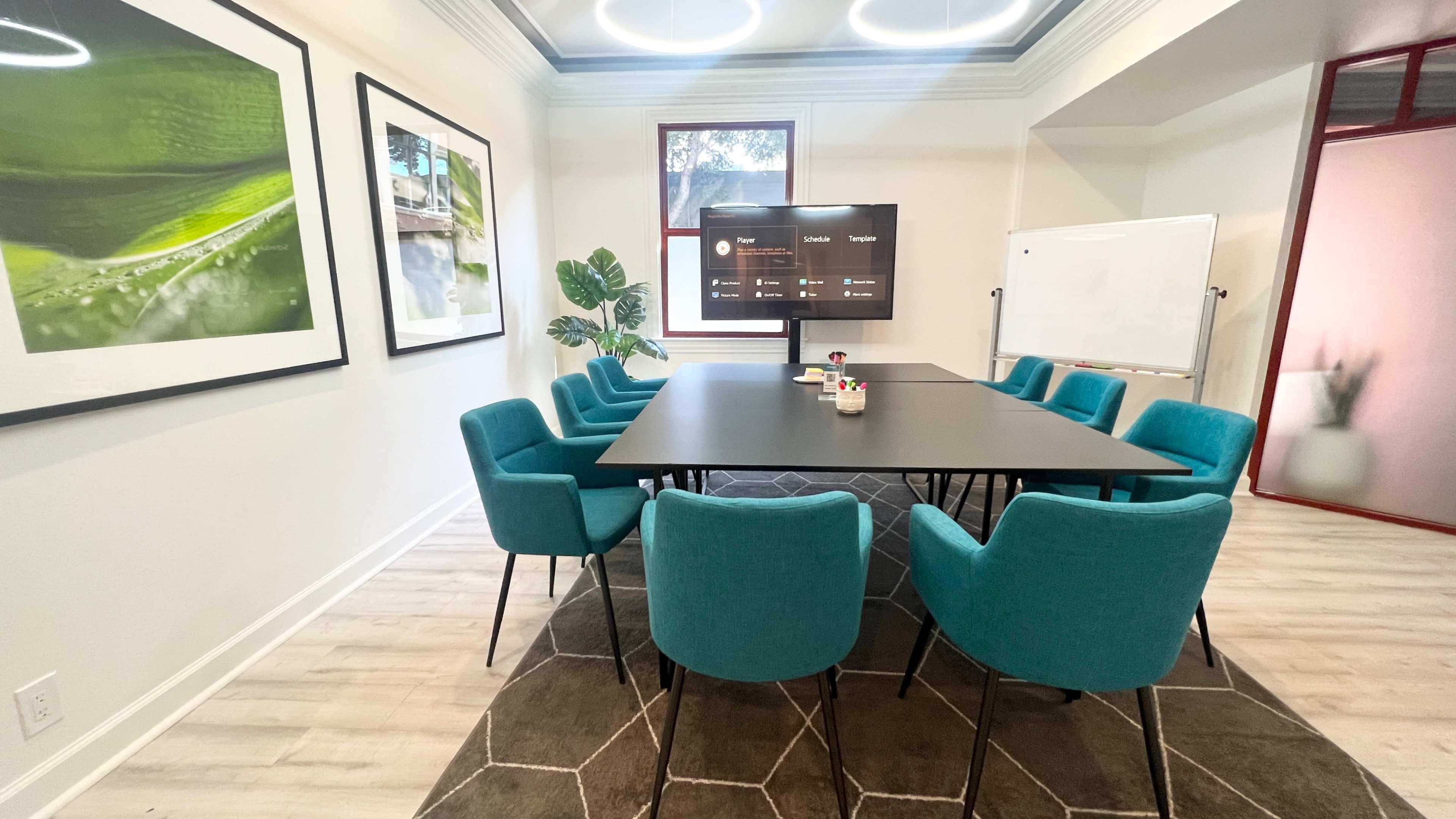 A meeting room features a large black table surrounded by teal chairs, a presentation screen, and framed nature photographs on the walls.