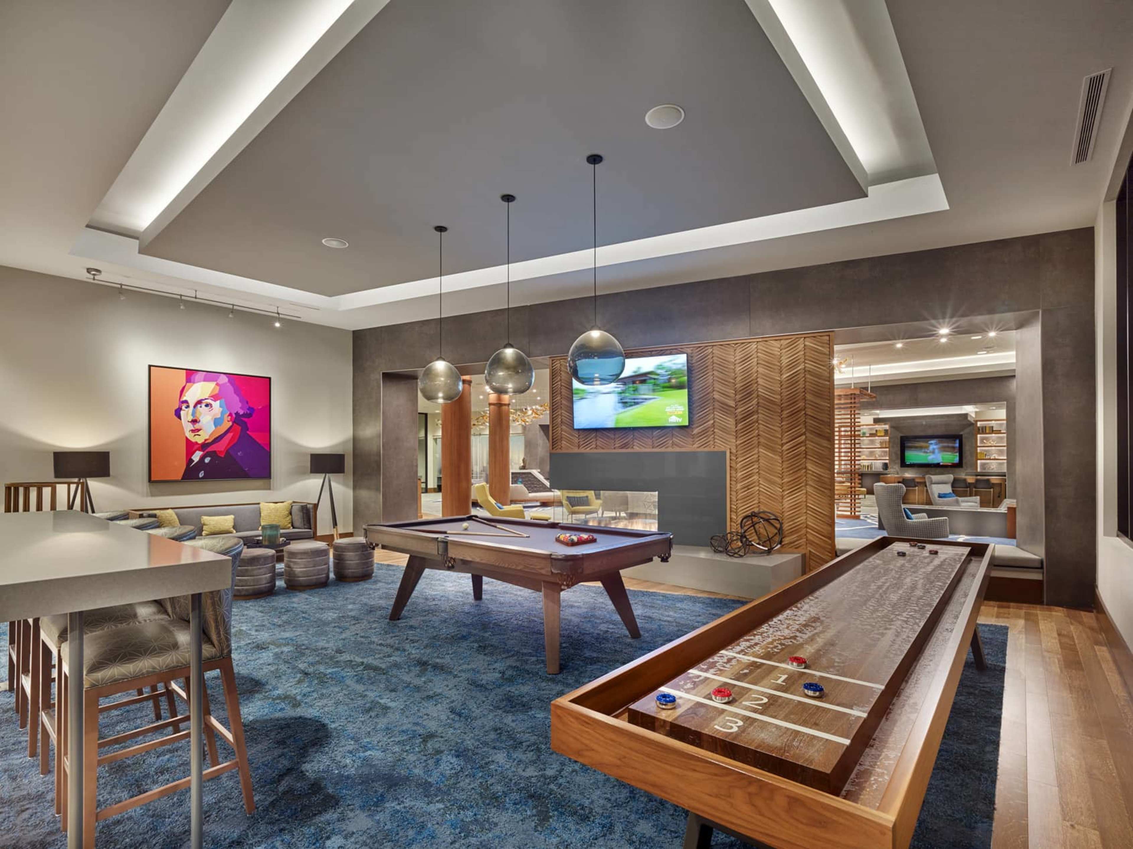 A modern game room with a pool table, shuffleboard, and an illuminated ceiling, featuring a colorful wall art piece and a television in the background.