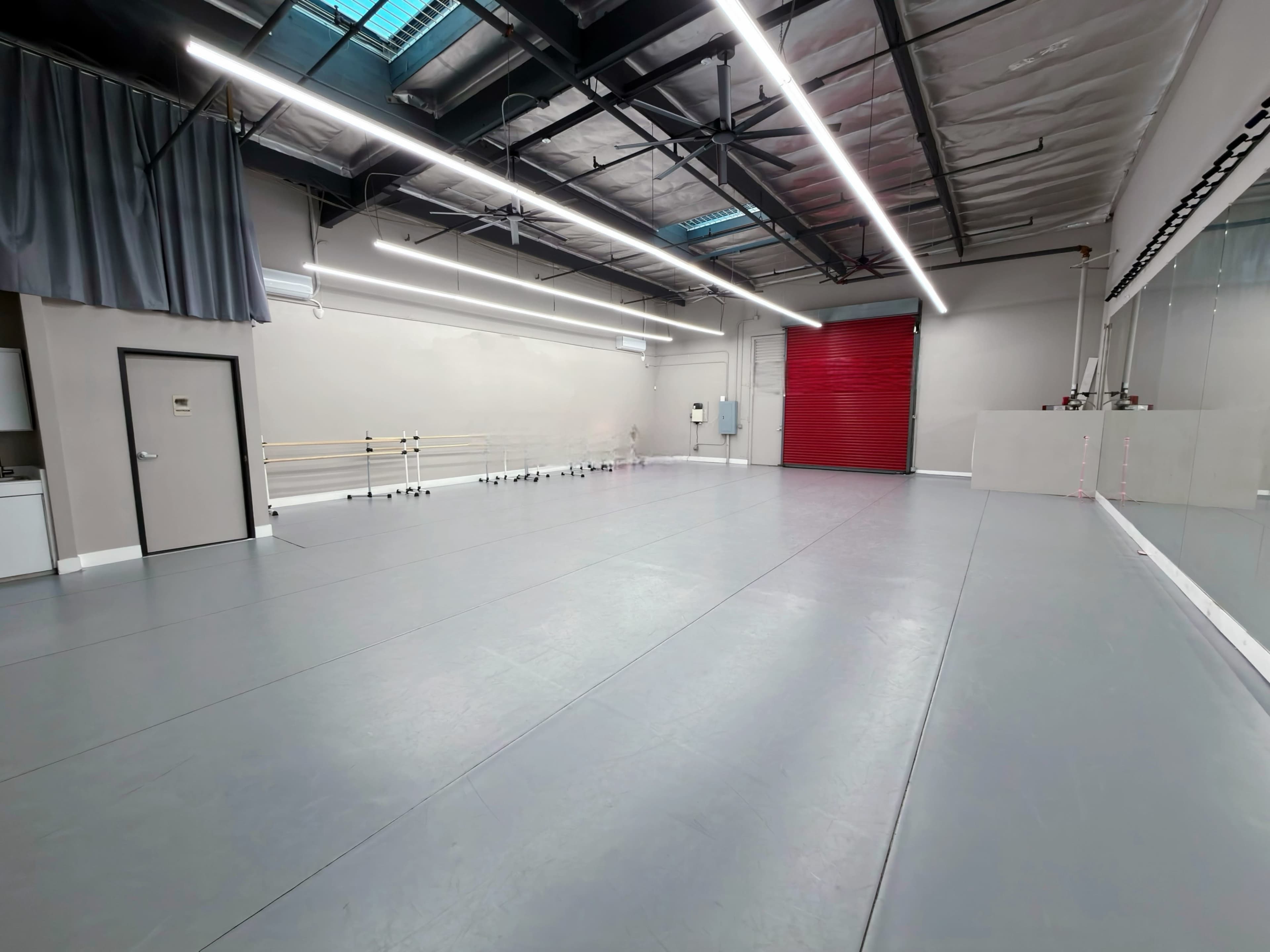 The image shows a spacious dance studio with gray flooring, mirrored walls, and a red garage door.