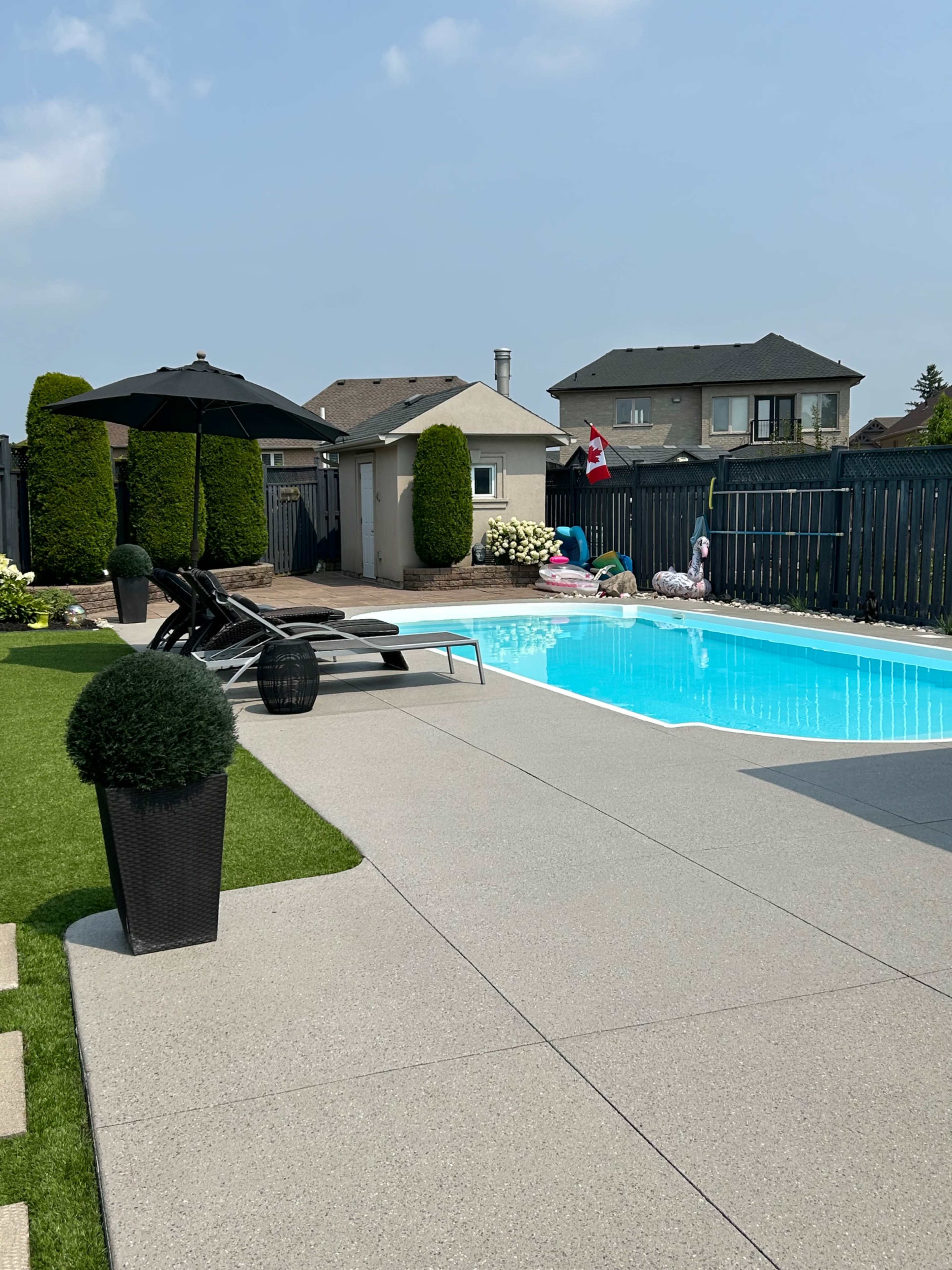 A backyard with a swimming pool, lounge chairs, and a patio area surrounded by fencing and greenery.