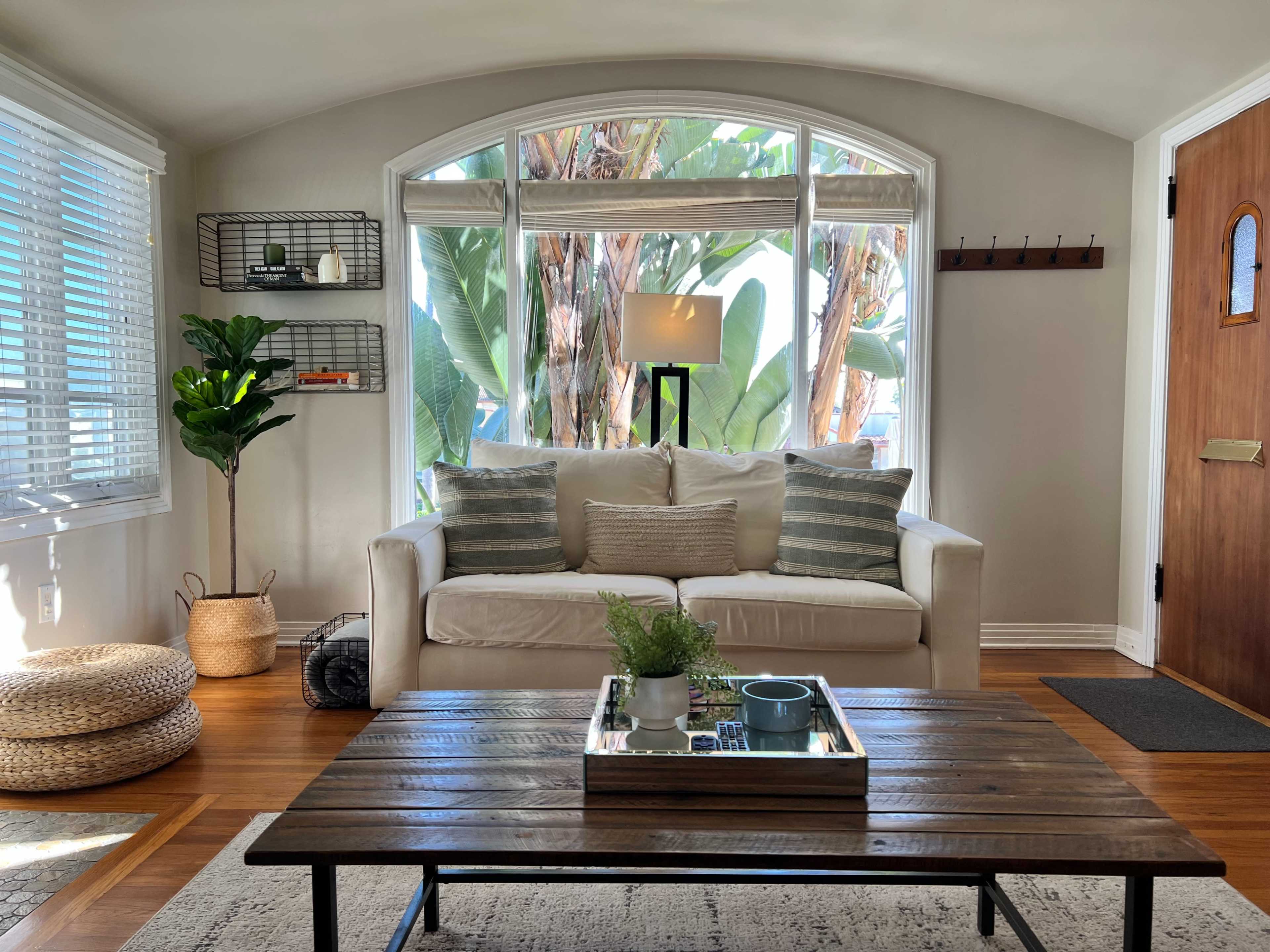 A well-lit living room features a light-colored sofa with cushions, a wooden coffee table, and large windows with views of palm trees.