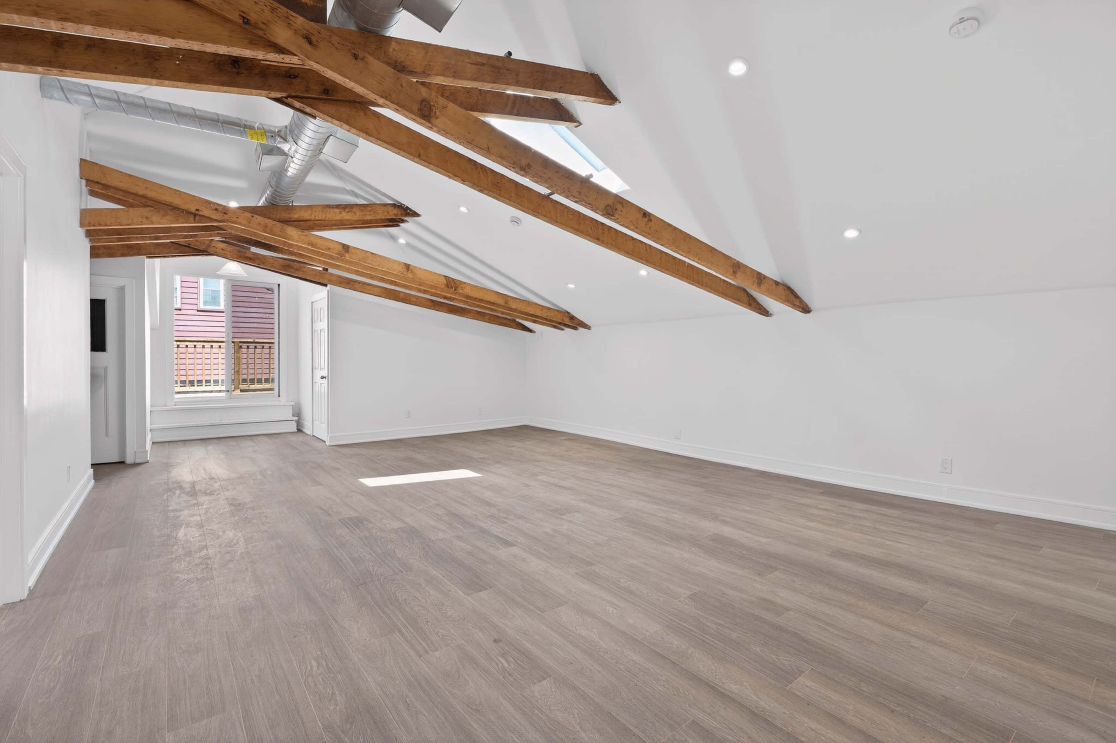 The image shows an empty room with wooden beams on the ceiling and large windows allowing natural light to enter.