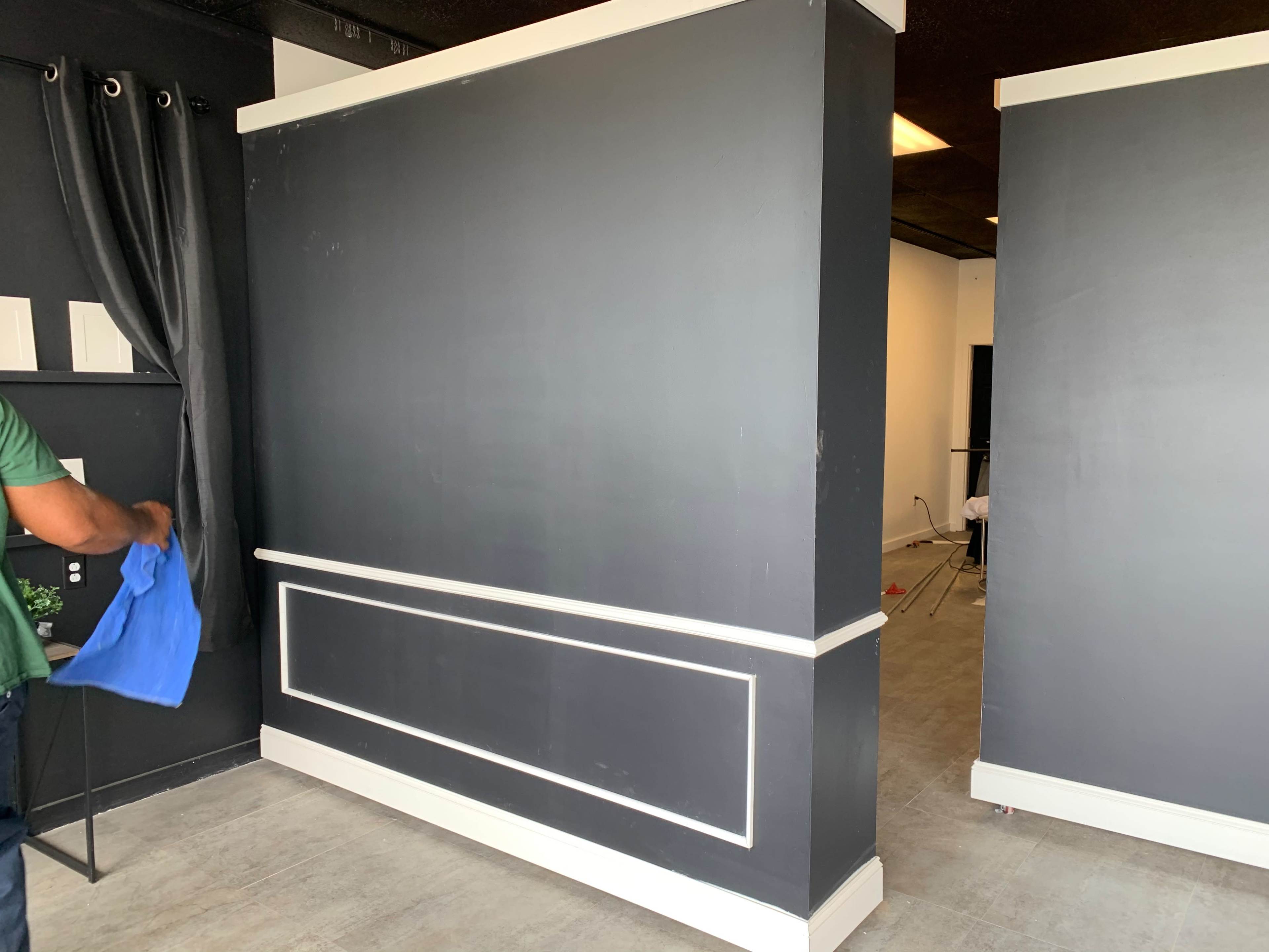 A person wearing a green shirt wipes a dark wall in a room with gray flooring and minimal furnishings.