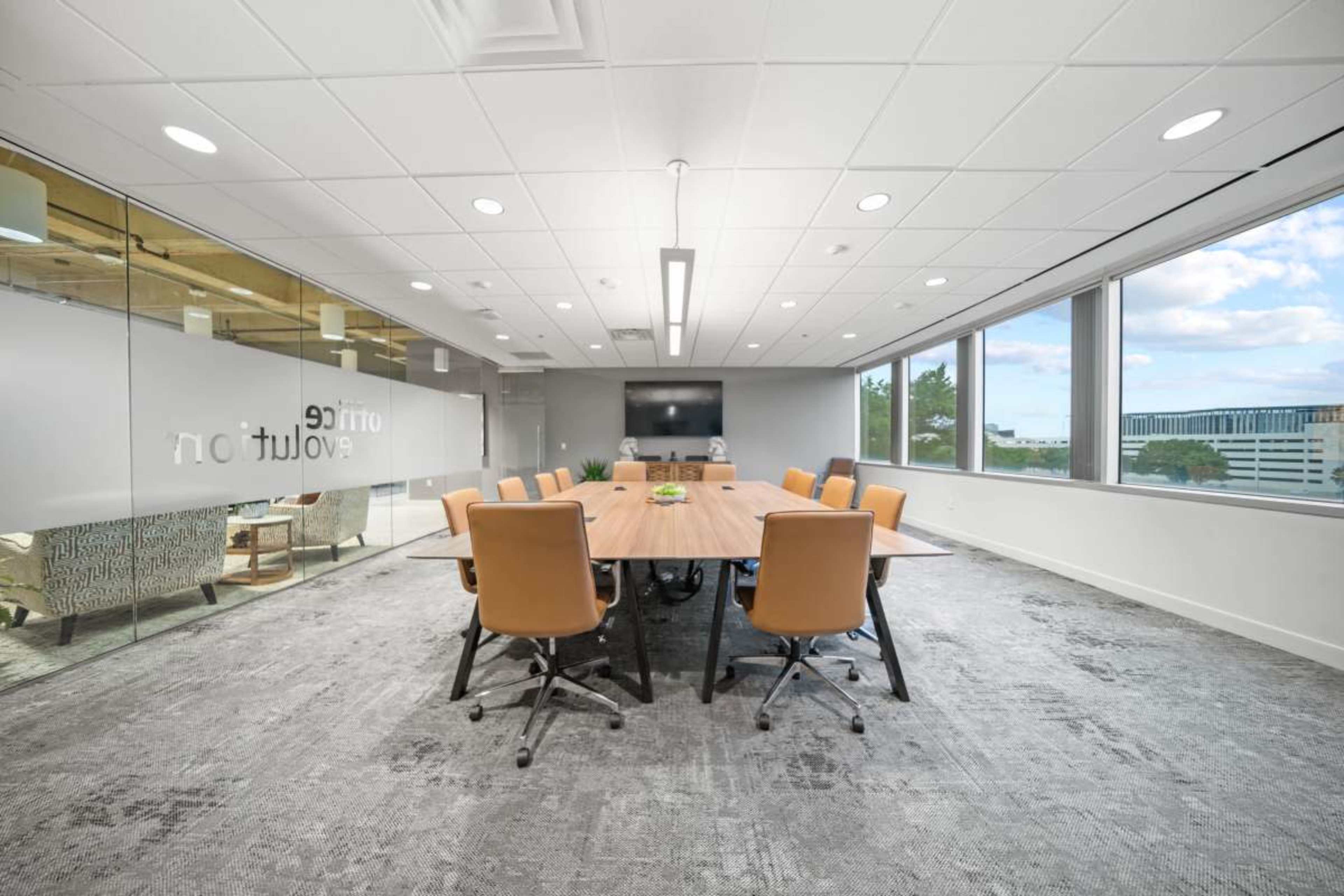A conference room features a large wooden table surrounded by orange chairs, with windows providing a view of the outside and a large screen mounted on the wall.