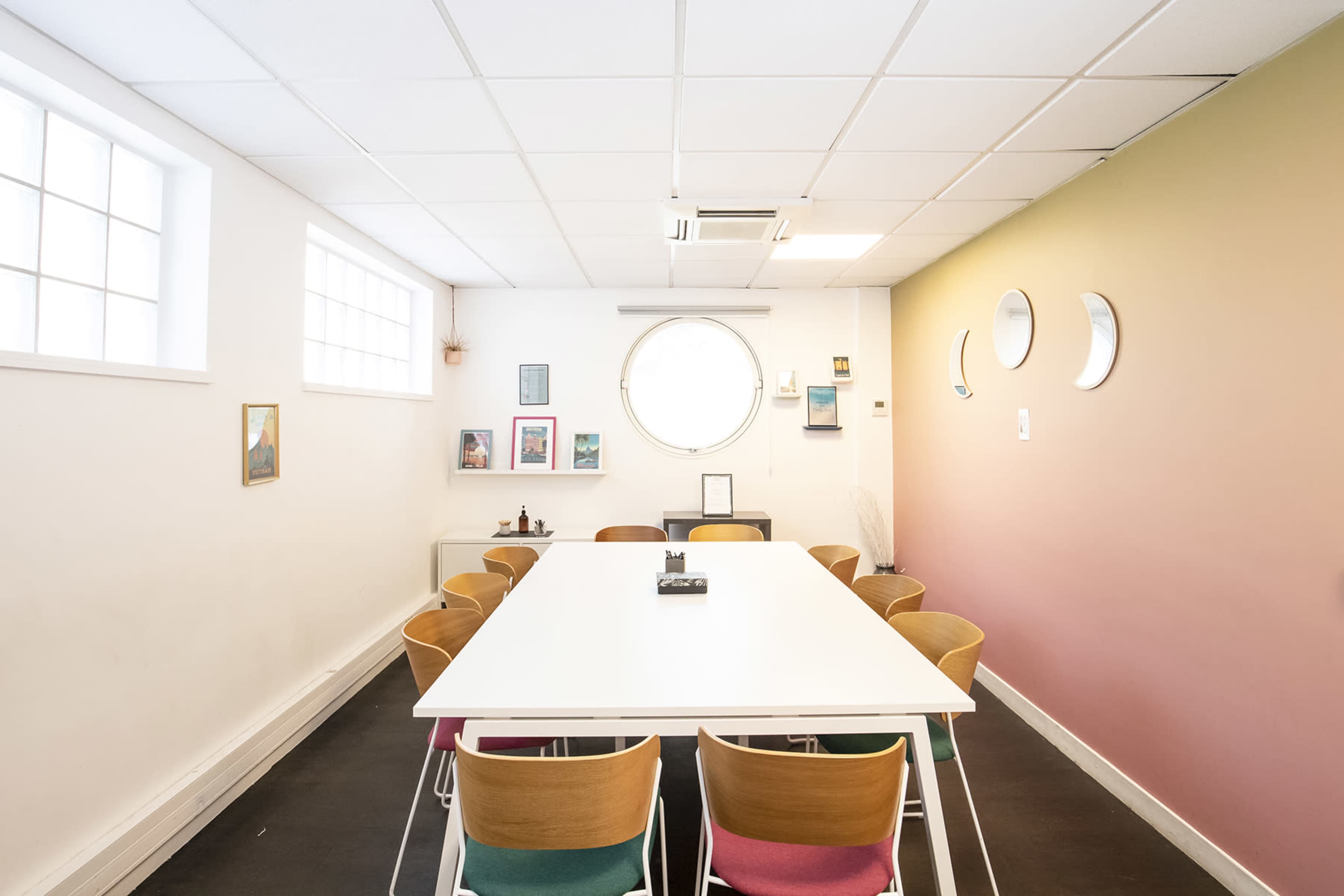 A bright, minimalist meeting room features a large white table surrounded by chairs, with framed artwork and a round window on the walls.