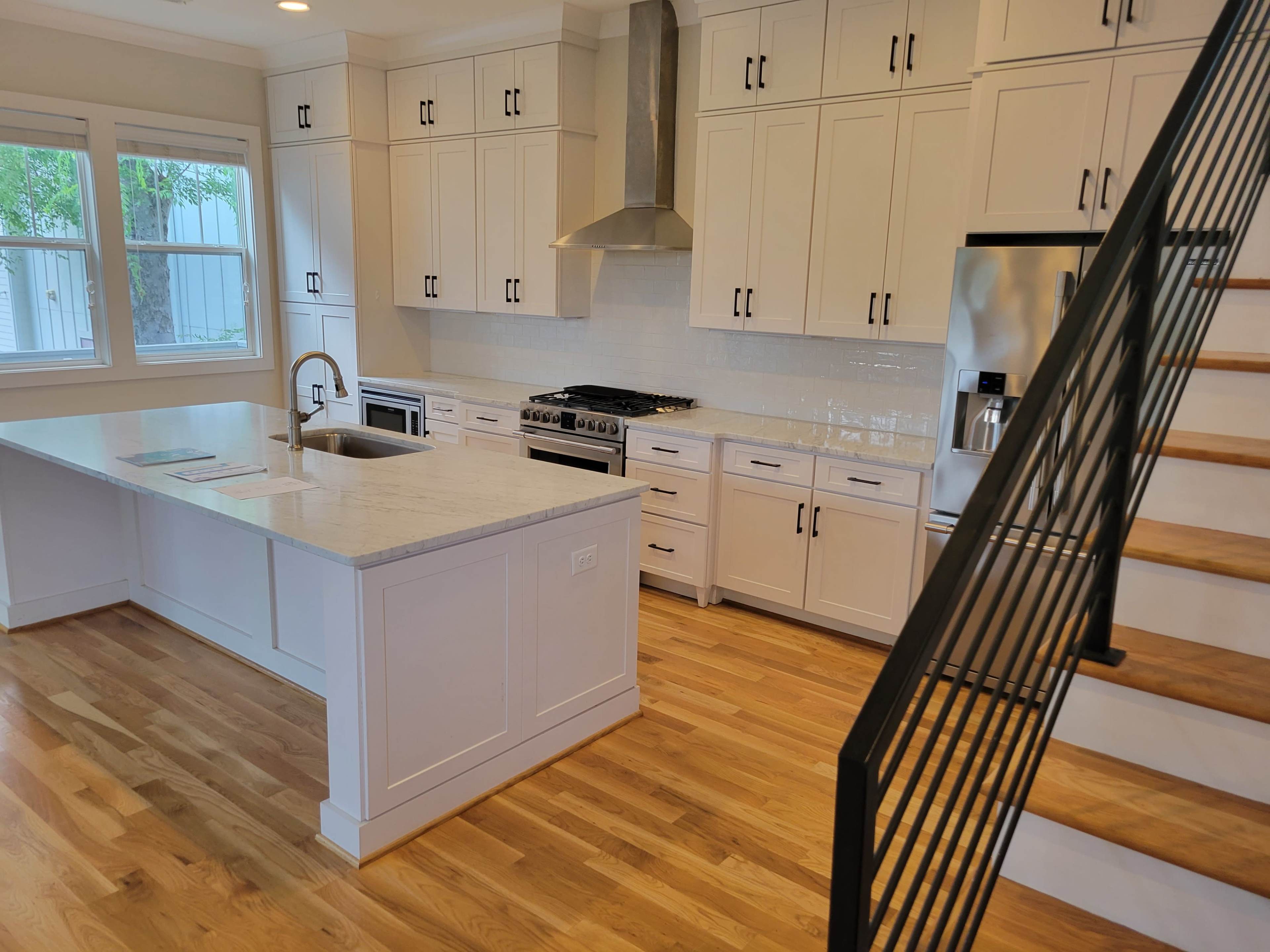 The image shows a modern kitchen with white cabinetry, a large island, stainless steel appliances, and a wooden staircase in the background.