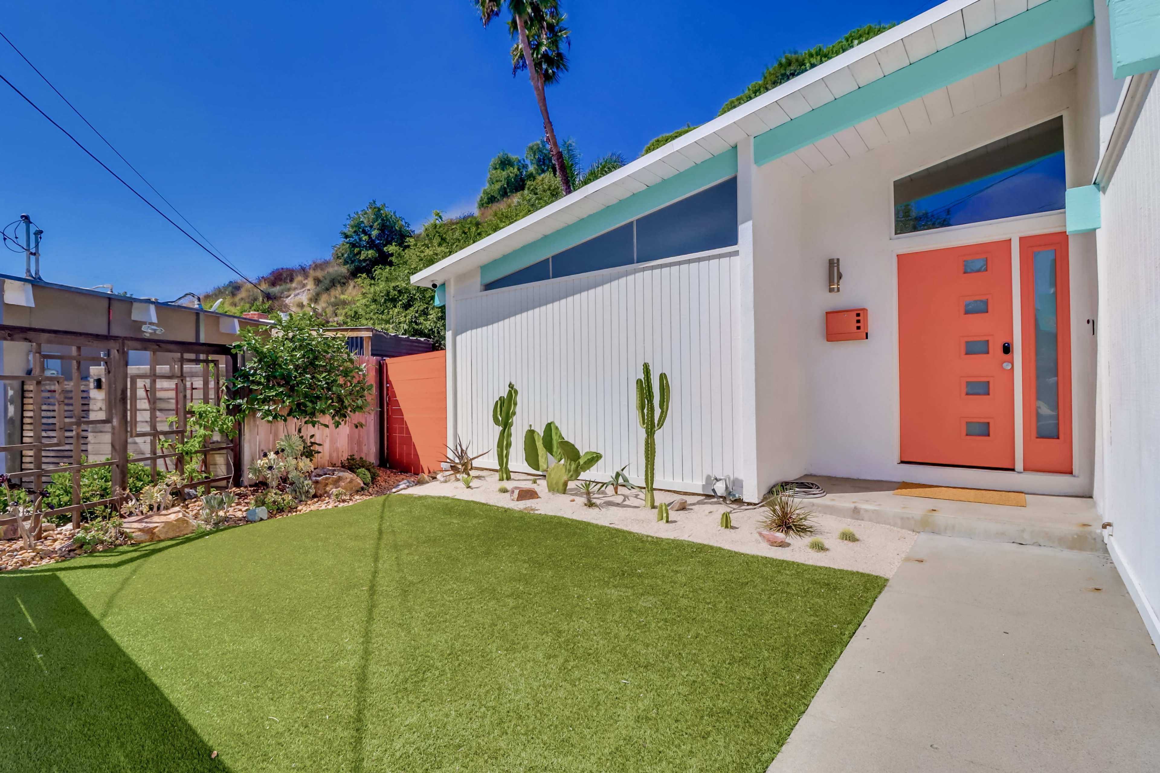 A modern house features a vibrant orange front door, surrounded by a landscaped yard with artificial grass and various cacti.