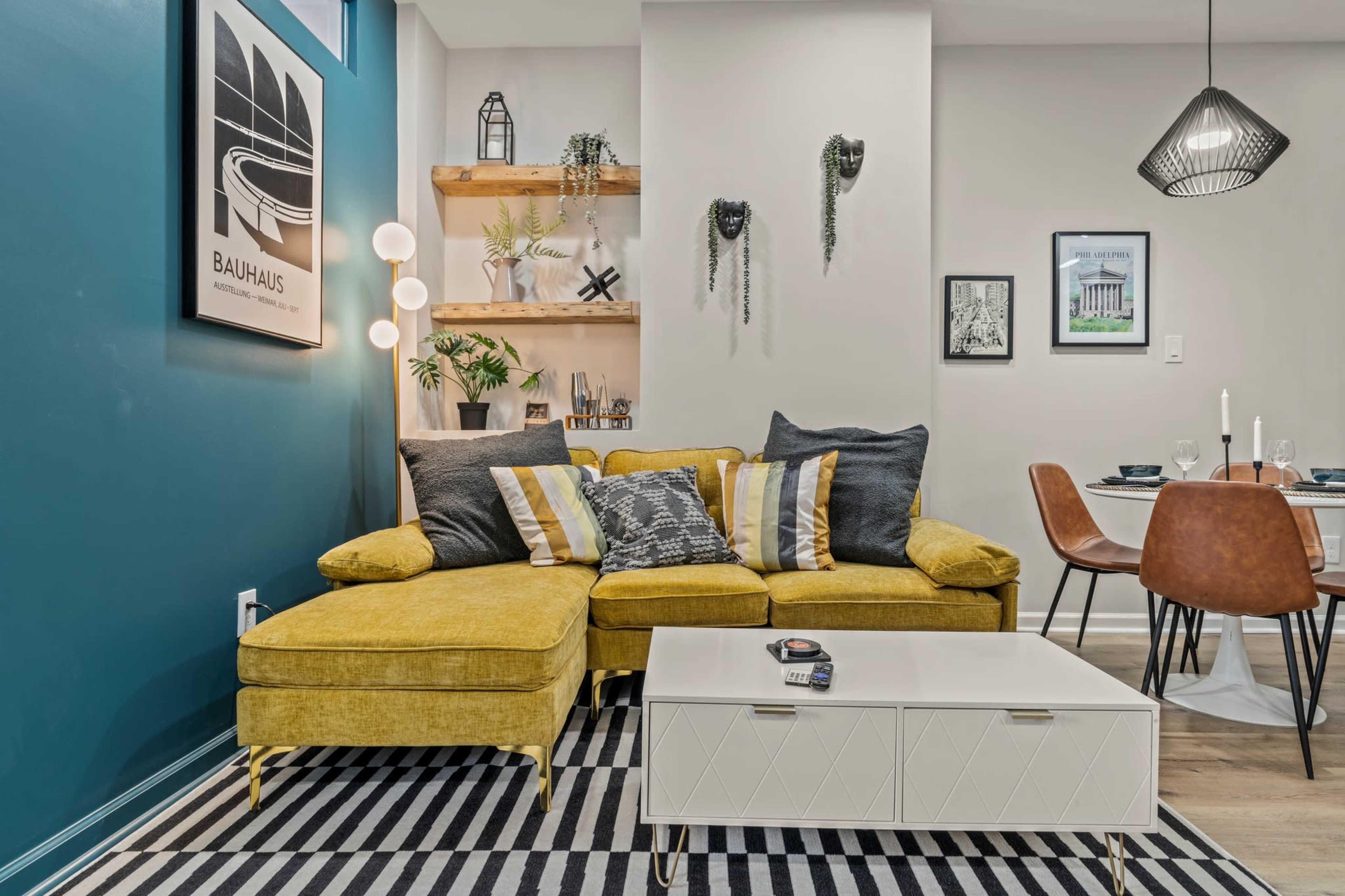 A yellow sectional sofa with decorative pillows is positioned in a modern living room characterized by a blue accent wall, a white coffee table, and a dining area in the background.