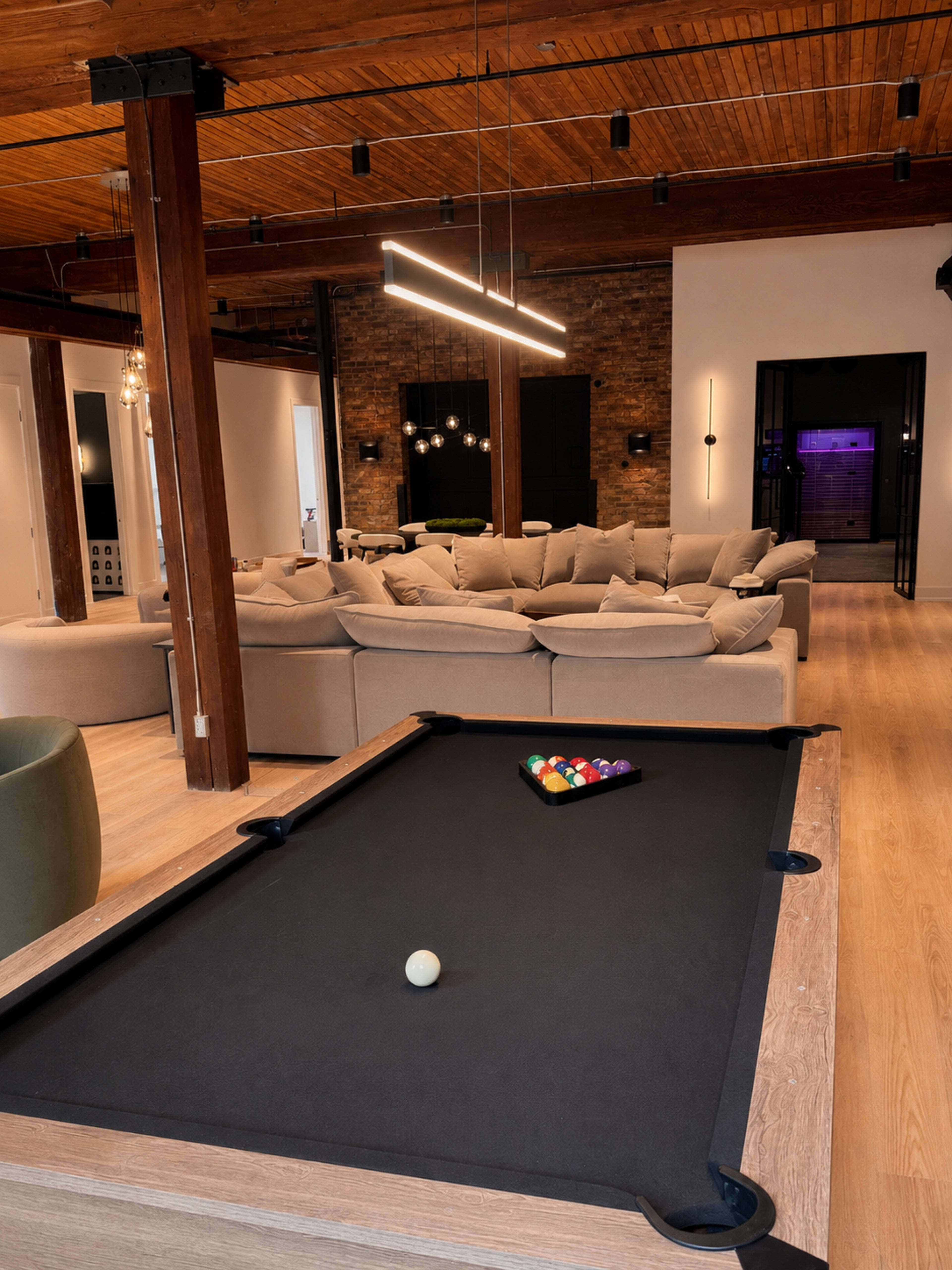 The image shows a modern living space featuring a pool table in the foreground and a large, cozy seating area in the background with warm wooden accents and soft lighting.
